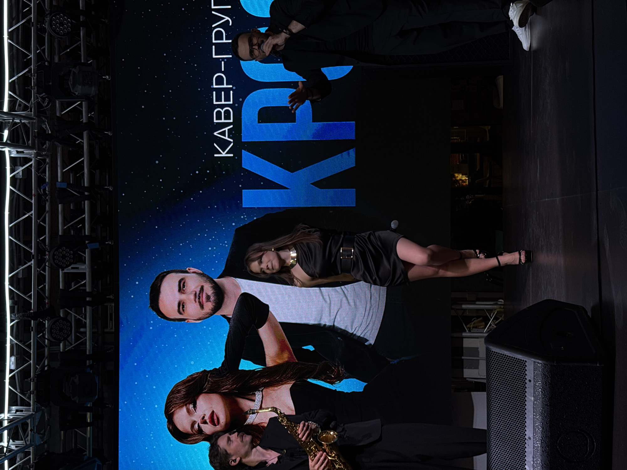 The image shows a promotional backdrop screen featuring a performance group, possibly in Moscow, Russia, based on the Russian text visible in the background which reads “КАВЕР-ГРУП”, meaning "Cover Band".
The backdrop features images of a man, a woman in a black dress with a gold necklace and belt, and a man playing a saxophone next to a woman with long hair. Another man wearing a suit is visible at the top of the screen.
Stage lighting equipment is visible on the left, suggesting a performance venue. A speaker is positioned to the right of the screen.
