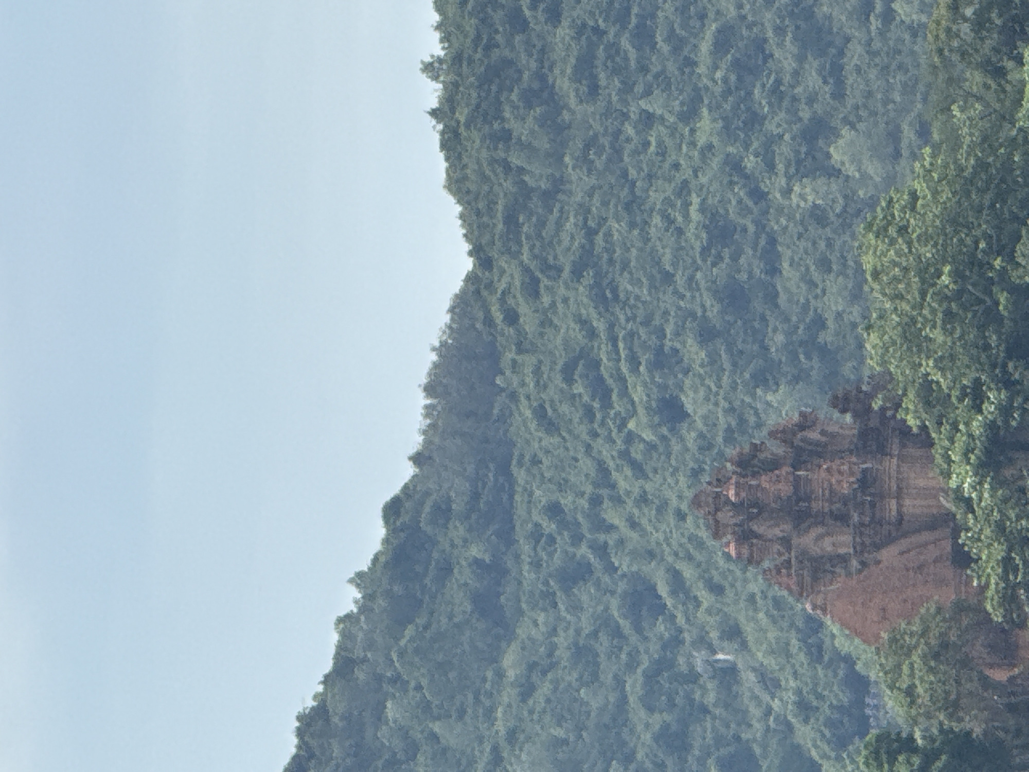 The image shows a mountain covered in dense green foliage, adjacent to a light blue sky. At the base of the mountain, nestled within the trees, is an ancient brick structure. The scene is presumably located in the Nha Trang Ward, Vietnam, as indicated in the prompt. The structure appears to be historical or archeological. No people, actions, or interactions are visible in the image.