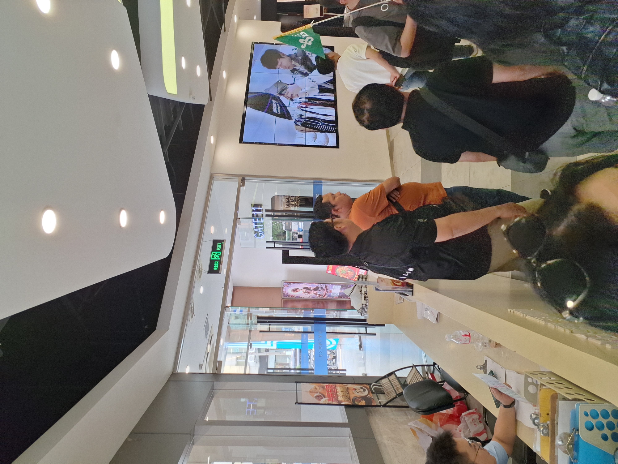 The image shows a group of people in what appears to be a commercial establishment, possibly a retail store or a mall, located in Hongkou District, China. A man in a light blue shirt is visible behind a counter, seemingly engaged with documents. Several other individuals are standing in a line or gathered in the foreground and midground, some looking towards a large screen displaying two individuals. A green flag with a symbol is held up by one person. The environment includes a counter with various items, a glass door entrance with signage, and a suspended ceiling with lighting fixtures. The overall scene suggests a public gathering or an event taking place.
