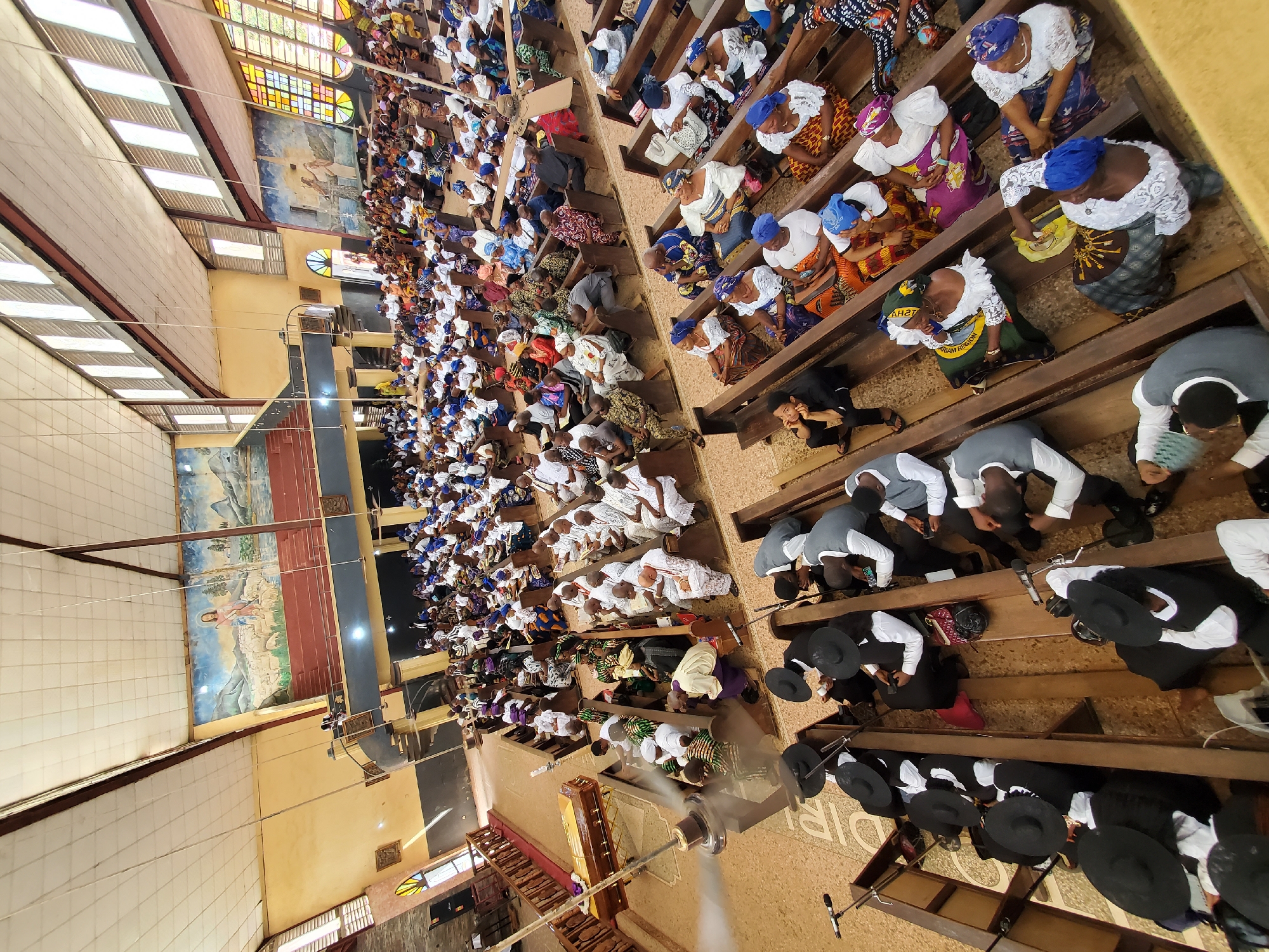 A high-angle, eye-level shot depicts a church service in Awkuzu, Nigeria. Rows of congregants, mostly women in vibrant traditional clothing and headwraps, fill wooden pews. Some individuals kneel in prayer while others sit. At the front, men in black suits and hats occupy the pews. An aisle leads to the altar area where a brown coffin is visible. Overhead, a ceiling fan rotates. Stained-glass windows and large murals adorn the church walls. The architecture includes a corrugated iron roof.
