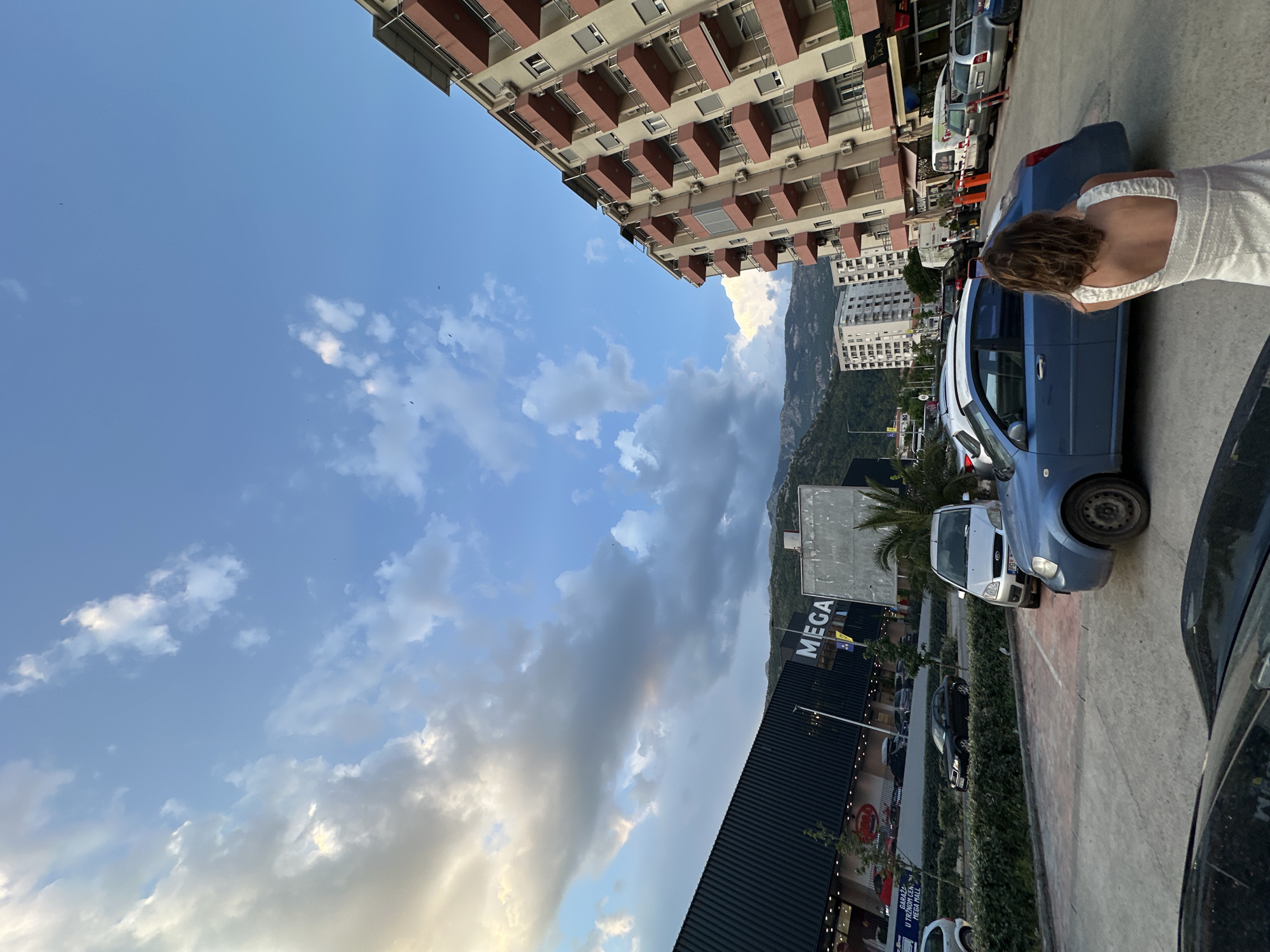 This media file captures a street scene in Budva, Montenegro. The image is oriented vertically, showing a view from a street level, looking upwards towards buildings and the sky. In the foreground, a woman wearing a white dress is seen from behind, standing near a blue car.
The scene appears to be set during the late afternoon or early evening, indicated by the lighting and the presence of clouds in the sky, some of which are illuminated by the setting sun. The weather seems to be pleasant, with a mix of clear blue sky and some dramatic cloud formations.
Several cars are parked along the street, and buildings line one side of the road. One prominent building in the background is a large, multi-story structure with a sign that reads "MEGA". Further in the distance, mountains are visible, suggesting a coastal or mountainous region. The overall atmosphere is that of a typical urban or commercial area during a day out.
The image is framed in a way that emphasizes the contrast between the urban environment and the natural landscape, with the sky and mountains forming a backdrop to the buildings and parked vehicles. The presence of the woman and the cars suggests a lively and active setting.