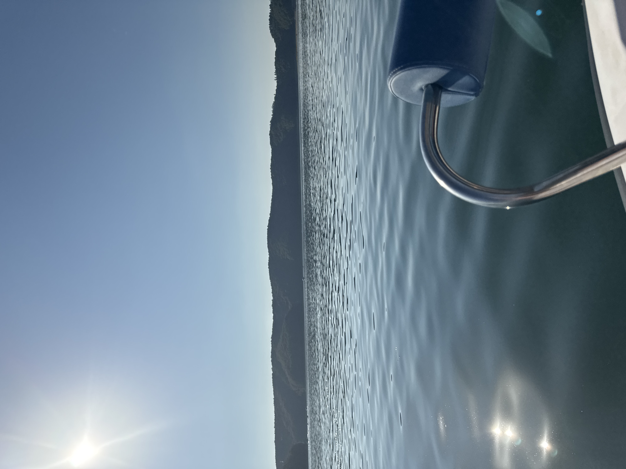 A wide-angle, eye-level shot captures a serene view of the San River in Polańczyk, Poland, on a bright, clear day. The sun, a brilliant white orb with distinct rays, shines from the upper left corner of the frame, casting a strong glare on the water. The river, a deep blue, is slightly rippled, reflecting the sunlight in shimmering patterns. In the foreground, a portion of a boat is visible, with a curved, chrome railing and a blue padded headrest. The distant shore is lined with lush, green, tree-covered hills, forming a gentle, undulating horizon against the pale blue sky. The overall impression is one of peaceful tranquility, with nature's beauty dominating the scene.
