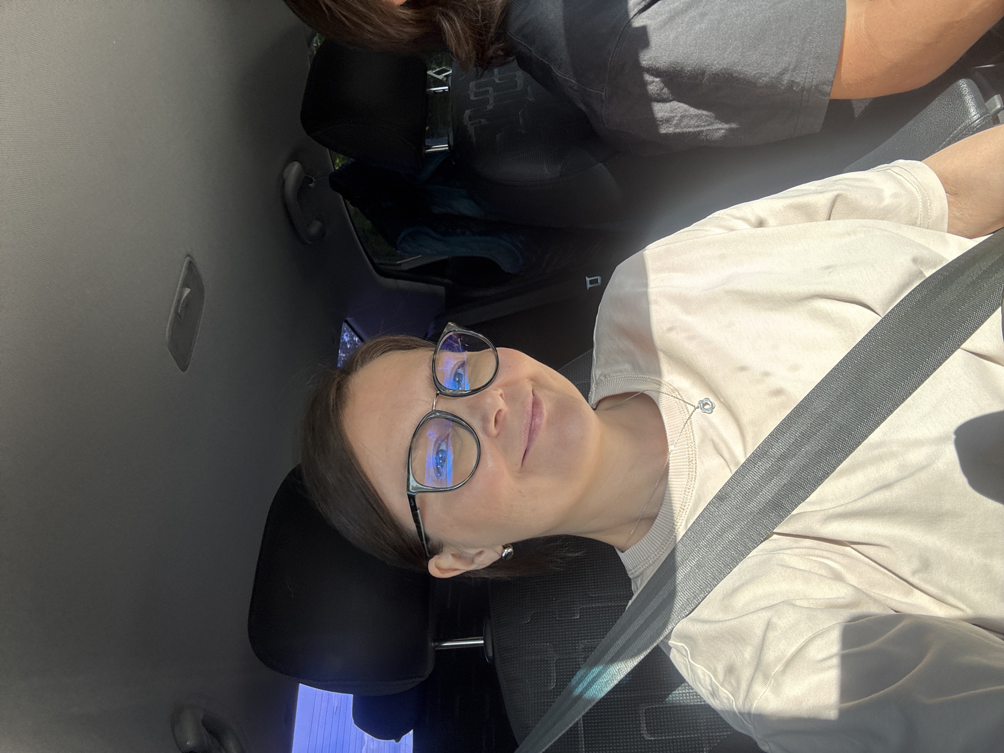 The image, oriented vertically with the main subject's head to the left, captures a woman in the front passenger seat of a car, smiling subtly. She has dark hair and wears black-framed glasses that show prominent blue reflections, possibly from blue light filtering. She is wearing a light-colored (beige or light grey) long-sleeved top and a thin silver necklace with a small, circular pendant. A grey seatbelt is securely fastened across her chest and lap.
Partially visible in the driver's seat is another individual, indicated by dark hair and a dark-colored top. The car's interior features a light grey fabric headliner, a grey grab handle, and an overhead light fixture. The seats are dark-colored. Bright sunlight streams into the vehicle, casting noticeable shadows on the woman and the interior, indicating a sunny daytime setting. Outside the car windows, blurred greenery and bright light can be seen. There is no visible text in the image. This scene takes place in Izhevsk, Russia.