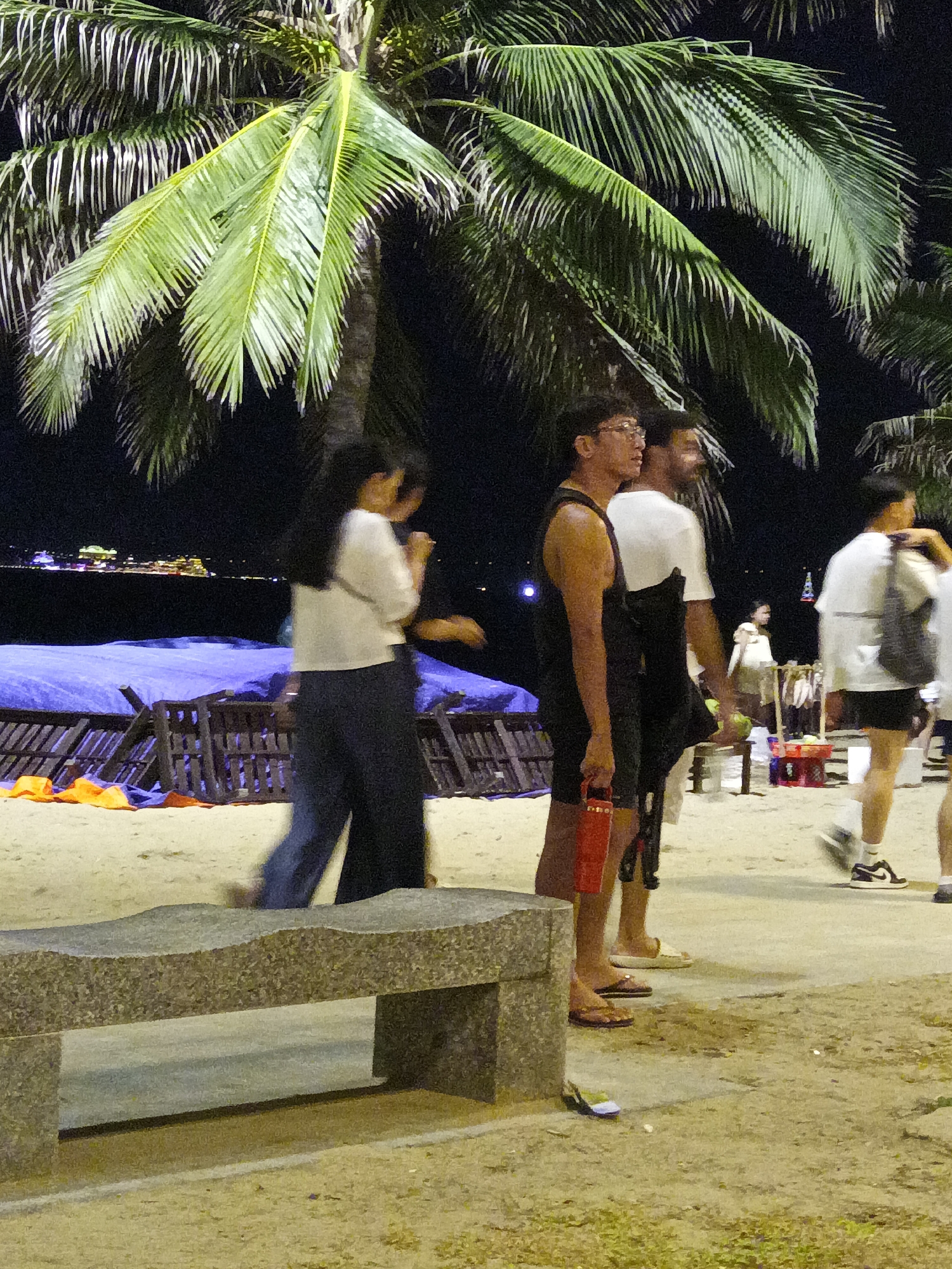 The image depicts a nighttime scene on a sandy beach, likely in Nha Trang, Vietnam, with a prominent palm tree dominating the left side. Several people are visible. In the foreground, a stone bench sits on the sand. To the right of the bench, three individuals are walking and standing. One person, seen from the side, wears a white t-shirt and dark pants, their long dark hair visible. Next to them, a person in a black tank top and black shorts holds a red cylindrical object and wears glasses. To their right, another person in a white t-shirt and black shorts walks away from the camera. Further in the background, other figures are present, along with beach furniture and blue tarps. The distant shoreline features illuminated buildings and lights, suggesting an urban backdrop. The lighting is artificial, creating a dimly lit atmosphere, with the palm fronds catching some of the light.