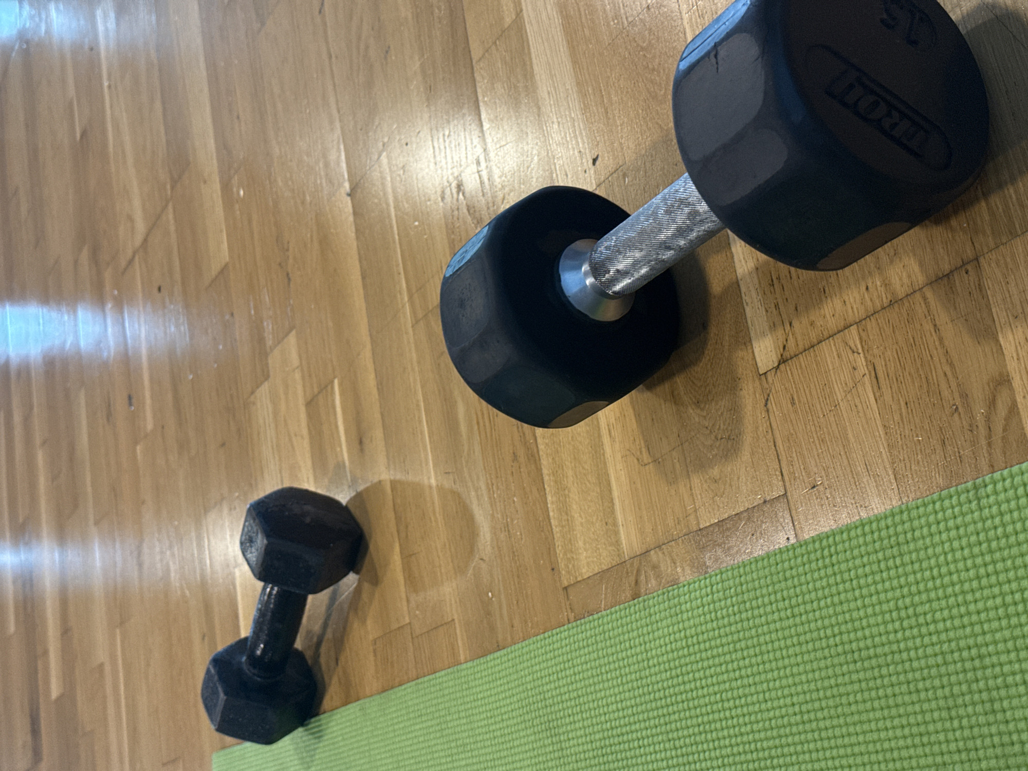 The scene presents two black dumbbells of different sizes resting on a wooden floor partially covered by a green yoga mat. The larger dumbbell, situated in the upper right quadrant, is labeled "IRON." The smaller dumbbell is positioned in the lower-left quadrant. The wooden floor consists of parallel wooden planks. The image suggests an exercise area. There is no evidence to ascertain that this image was taken in Mexico City, Mexico.