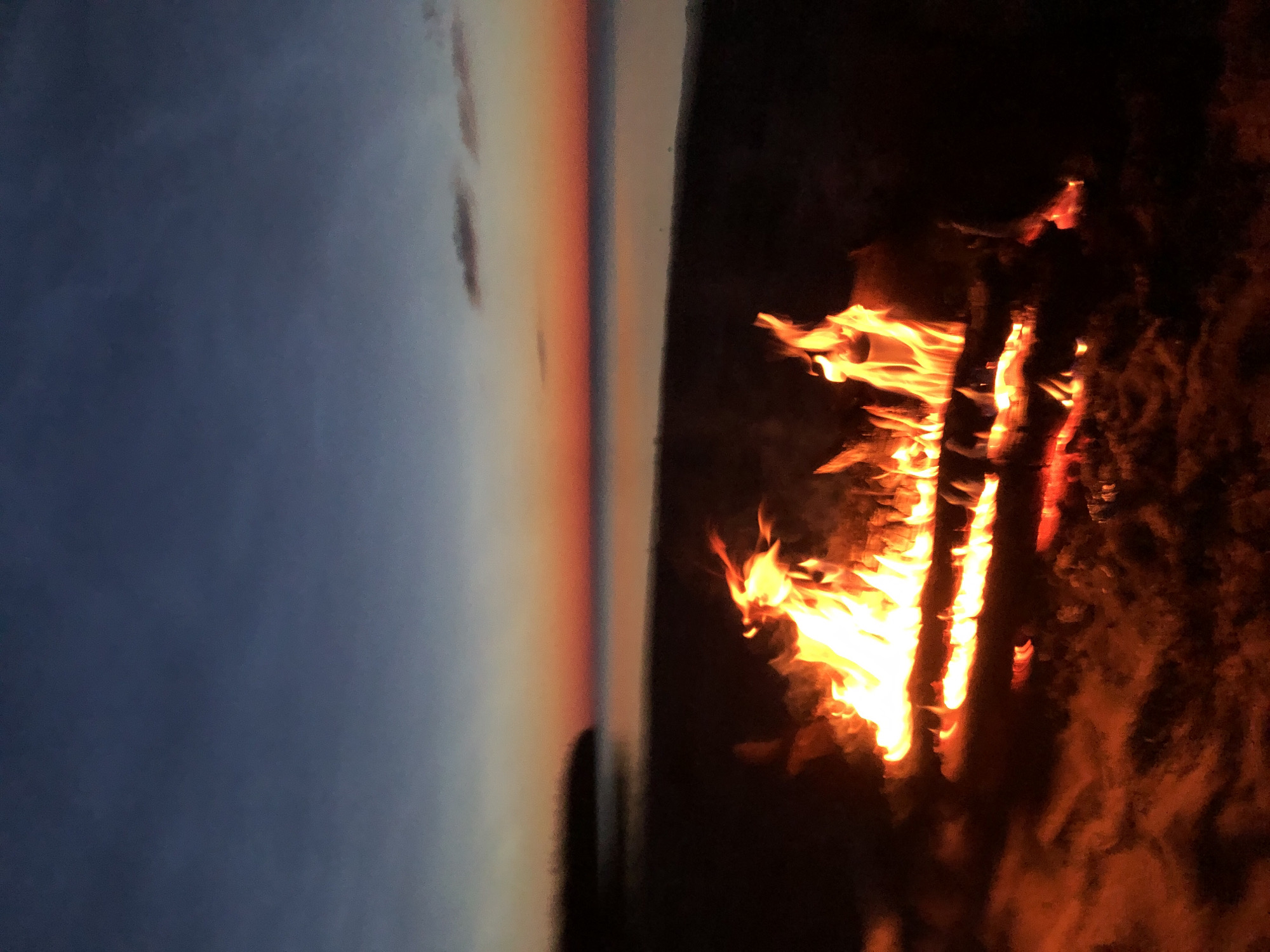 A bonfire burns on a beach at dusk. The sky is a gradient of deep blue at the top, transitioning to orange and pink at the horizon where it meets a body of water. A dark, indistinct landmass is visible on the left side of the image. The fire is composed of several logs, with bright orange and yellow flames licking upwards. The sand around the fire appears dark and damp. The overall scene suggests a tranquil evening by the water.