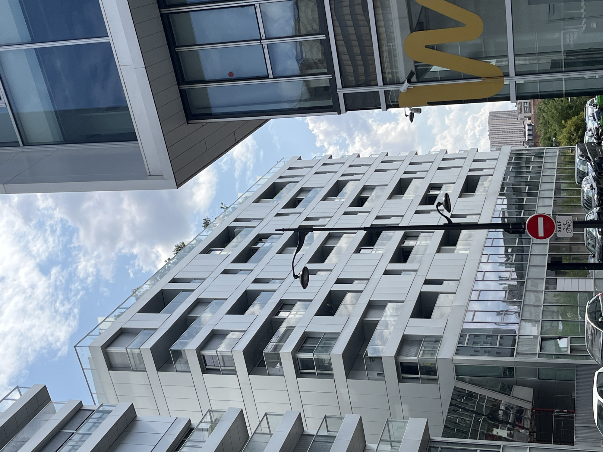 In a medium shot, two modern buildings with glass facades and balconies are captured against a cloudy sky. The building on the left is white with a layered design, featuring numerous balconies with glass railings. The building on the right is also modern, with large glass windows and a slanted, white overhang. A yellow, wavy logo is visible on the glass facade of the building on the right. A street lamp with a curved arm stands to the left of the building on the left. A red and white circular traffic sign with a horizontal bar indicates "Interdit" (forbidden) is mounted on a pole in front of the building on the left. Several cars are parked on the street in front of the buildings. In the background, other buildings and trees can be seen. The scene suggests an urban environment with contemporary architecture.