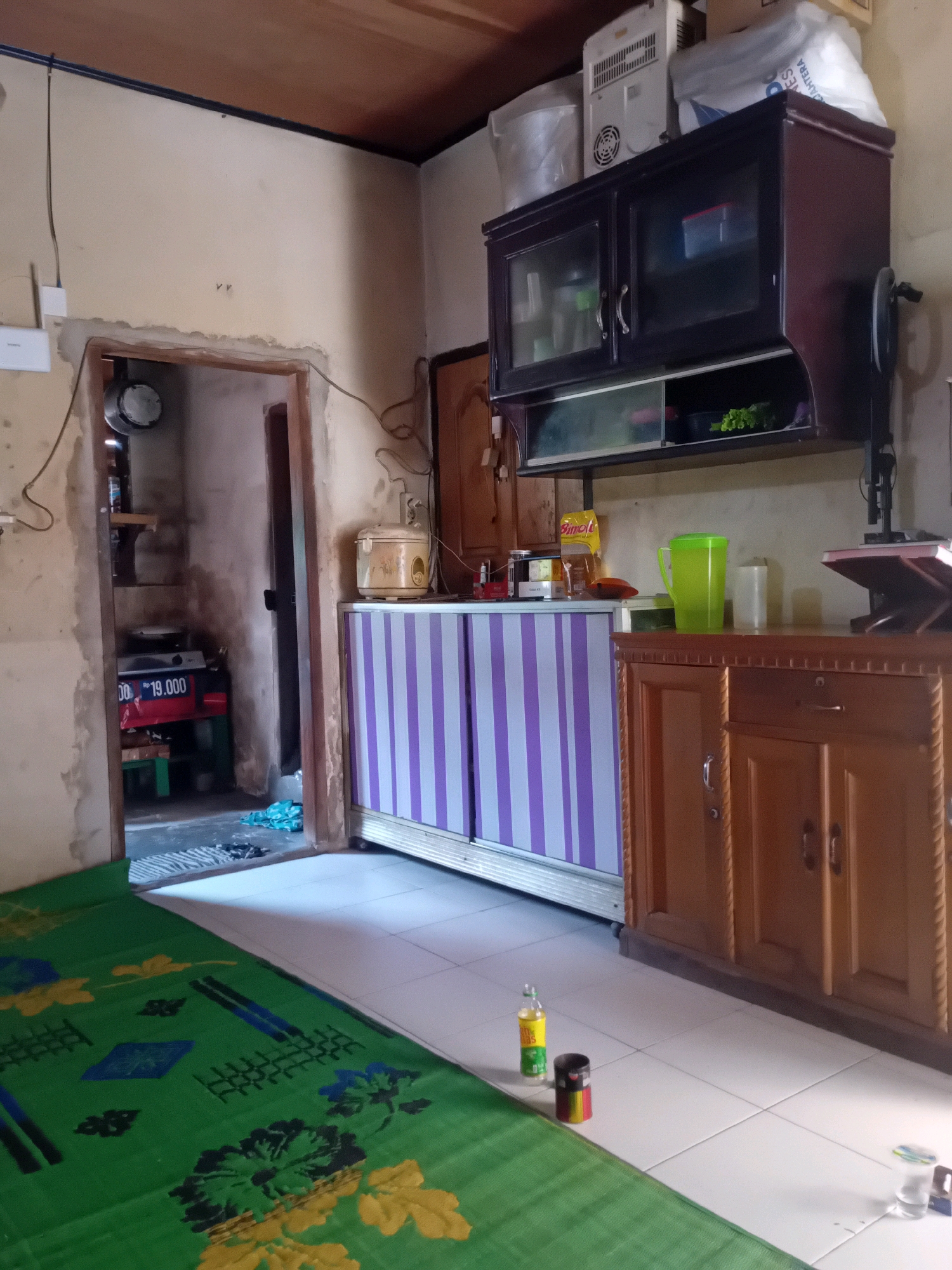 The image depicts the interior of a modest room, likely a kitchen or living area, with elements suggesting it is located in Pekanbaru, Indonesia. The room has light-colored, textured walls and a wooden ceiling. To the left, an open doorway leads into another dimly lit space where a gas stove and cooking utensils are visible, along with a price tag indicating "Rp 19.000". A green sleeping mat with a floral pattern covers the floor in the foreground.
Dominating the center and right of the image is a dark wooden cabinet mounted on the wall, featuring two glass-fronted doors on the upper section, displaying various items inside. Below this, a shelf holds a green pitcher, a white cup, and a small green plant. To the right of the cabinet is a wooden chest of drawers with metal handles and decorative carving.
In front of the wall cabinet is a low cabinet with alternating purple and white striped doors. A cream-colored rice cooker sits on top of this cabinet. On the floor, in front of the striped cabinet and to the left of the wooden chest of drawers, are a few small bottles and containers. The tiled floor extends from the foreground into the midground. The overall impression is of a lived-in, functional space.