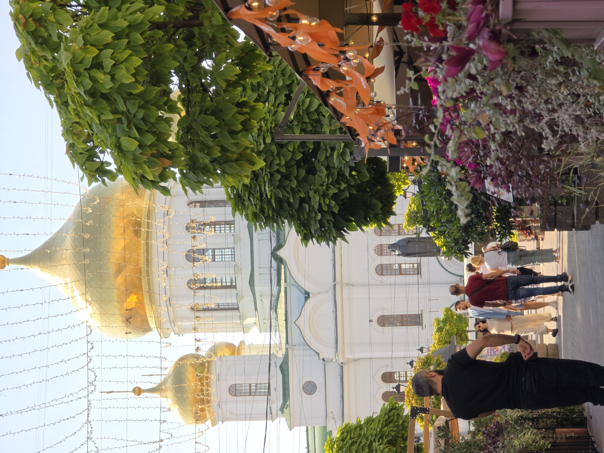 The image depicts a sunlit street scene in Rostov-on-Don, Russia, with a prominent white church featuring golden domes in the background. The church's architecture suggests it is the Rostov Savior Transfiguration Cathedral. In the foreground, a man in a black t-shirt and pants walks away from the camera, his back turned. Other people, dressed in casual attire, are walking in the same direction further down the street. The scene is bordered by lush green trees and vibrant flowering plants in planters, with decorative orange flags and string lights hanging above an outdoor seating area, possibly a cafe or restaurant. The overall atmosphere suggests a pleasant day in a city with a rich architectural heritage.