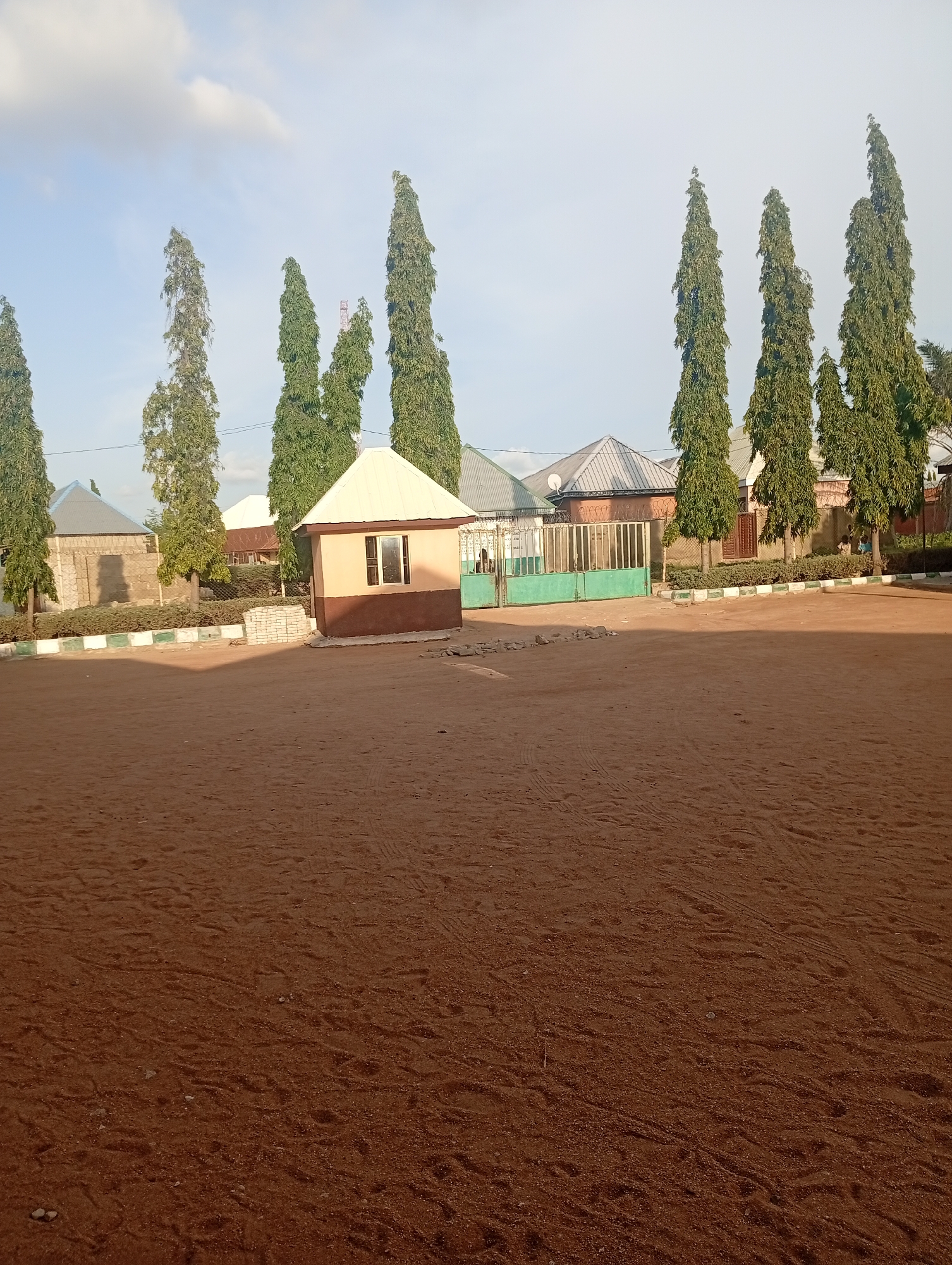 The image depicts an outdoor scene in Jalingo, Nigeria, likely a school or administrative compound. A large, open area of reddish-brown dirt occupies the foreground, with tire tracks and footprints visible. Along the back of the dirt area, a low border with white and green stripes runs horizontally. Behind this border, several buildings and structures are visible.
To the right, a small, octagonal building with a light-colored, sloped roof and brown lower walls stands prominently. It has a single window with dark framing and glass. Further to the right, a green metal gate is visible, partially obscured by a tall, slender cypress tree. Behind the gate, a fence and more buildings can be seen, including what appears to be a residential structure with a corrugated metal roof.
To the left of the octagonal building, a fence made of stone blocks and wire mesh runs across the mid-ground, separating the dirt area from a row of buildings with peaked, grey corrugated metal roofs. Several tall, slender cypress trees are planted in a line along the mid-ground, interspersed with these buildings. One building on the far left has a light blue, peaked roof.
The sky above is a clear, pale blue with scattered white clouds. The lighting suggests it is either early morning or late afternoon, with the sun casting shadows from the trees and structures onto the dirt ground. No people are clearly visible or engaged in specific actions within the frame. The overall impression is one of a functional outdoor space within a developed area, characterized by its dry, earthy terrain and the presence of several buildings and tall evergreen trees.