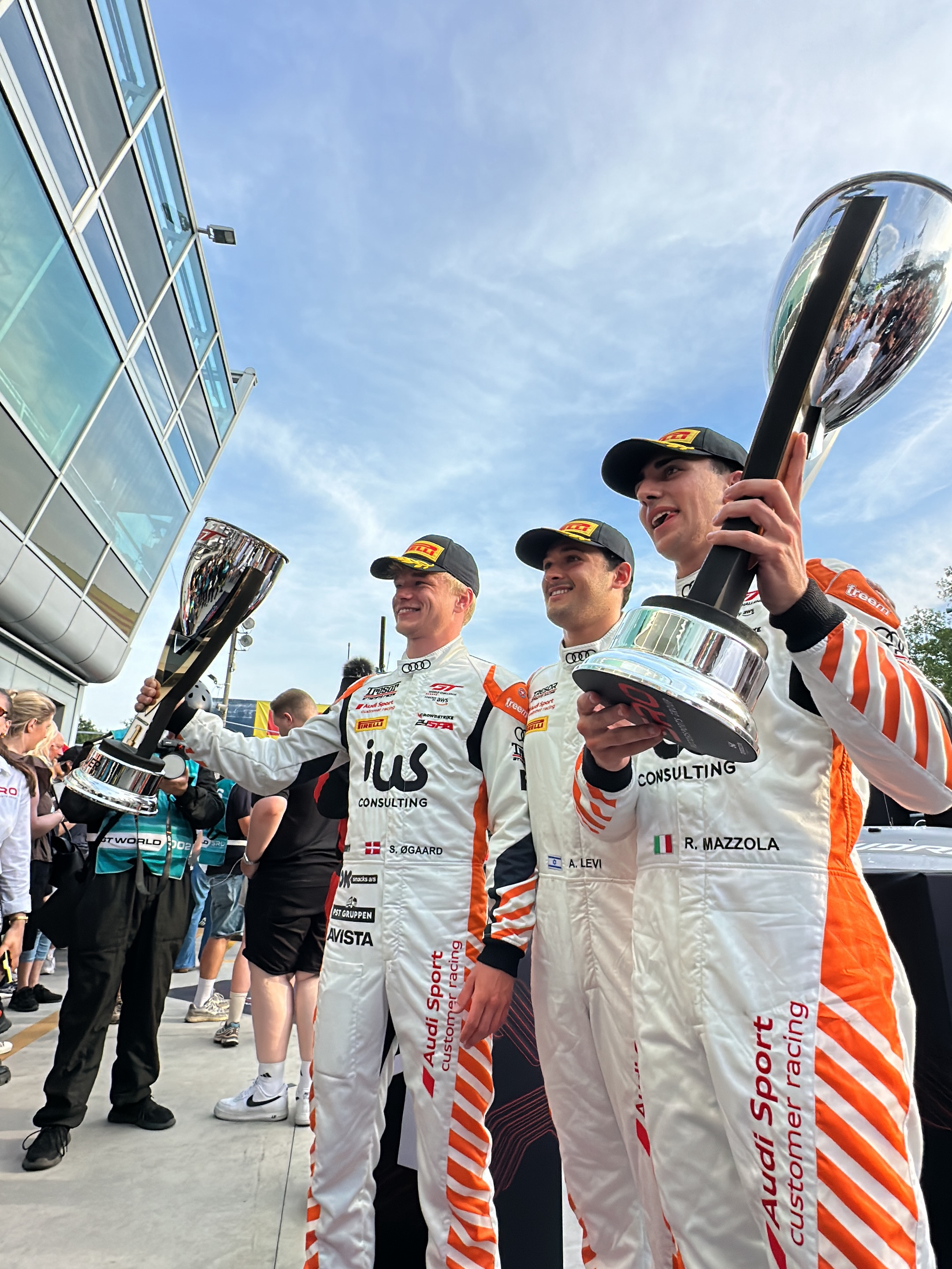 Three men stand close together, each holding a large, reflective trophy above their heads. They are dressed in white racing suits with orange diagonal stripes. Each suit has various sponsor logos on the front, including "ius CONSULTING" and "Audi Sport customer racing." Each suit also has the name of each person "S. ØGAARD", "A. LEVI", "R. MAZZOLA". The man on the left has a Danish flag on his suit, the man in the middle has an Israeli flag on his suit, and the man on the right has an Italian flag on his suit. All three men wear black caps with the PIRELLI logo. The background shows a modern, glass-walled building and a crowd of people, suggesting this is an outdoor event at a racing track. The sky is partly cloudy, giving the scene bright, natural lighting. The image appears to be taken in the city of Monza, Italy, where the Monza racetrack is located, and the suits could be from a motorsports event at the Audi Sport customer racing series.