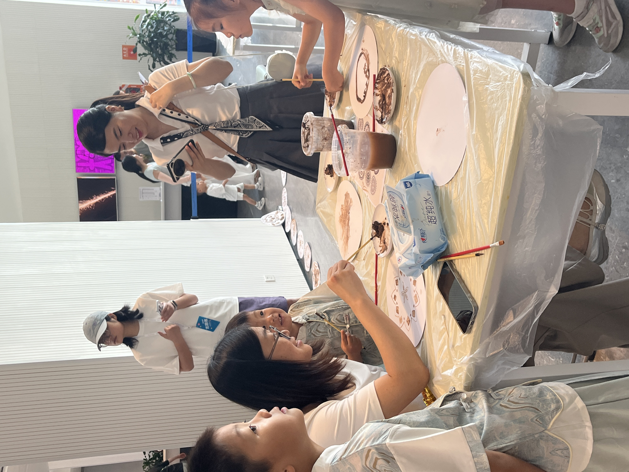 In an indoor setting that appears to be a public event or workshop, likely in the Chongqing High-tech Zone, China, a group of individuals, predominantly children, are engaged in a painting activity. A long table covered with a clear plastic tablecloth is the central focus, on which lie paper plates with designs, cups containing brown paint, brushes, and wet wipes.
Several children are seated around the table, actively participating. One girl, wearing a light green dress, is using a brush to paint a design on a paper plate. Another child, a young boy, is looking intently at his work. A woman, possibly an instructor or guardian, sits with the children, holding a brush and guiding them. She is wearing glasses and a white top. Another woman, dressed in a white shirt and a dark skirt, stands at the back, holding a smartphone and looking at it. In the background, another woman in a white t-shirt and a grey cap is also visible, holding a phone.
The environment suggests a bright, modern space with white walls and vertical paneling. A plant can be seen in the background, and digital screens displaying images are mounted on the wall. The overall scene depicts a hands-on, educational, or recreational activity for children.
