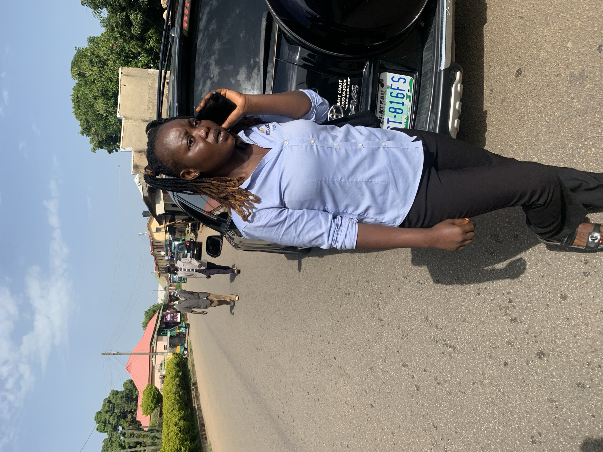 A dark-skinned woman with dreadlocks styled in pigtails is standing on a paved road next to a black SUV. She is wearing a light blue button-down shirt and black pants, and is speaking on a dark smartphone held to her right ear. The SUV has a license plate that reads "PLATEAU ST-816FS". In the background, two men in casual attire are walking away from the camera down the road. Further in the distance, there are buildings and trees, suggesting an urban or suburban environment. The sky is clear with scattered clouds. The overall scene appears to be in Jos, Nigeria, based on the "PLATEAU" license plate.