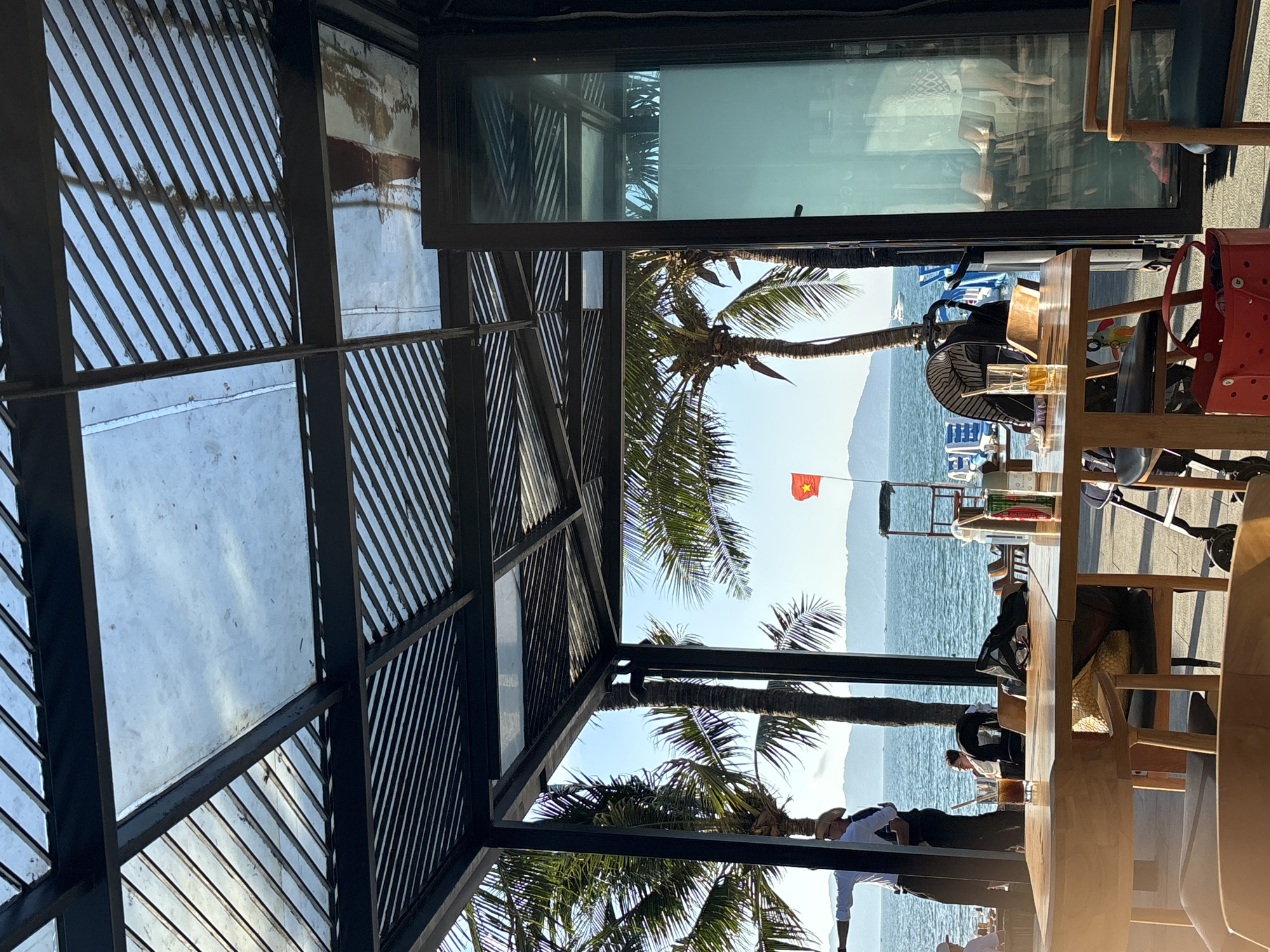 The image is taken from under a shaded structure, possibly a veranda or gazebo, in Đà Nẵng, Vietnam. Looking out through the structure's latticed roof, palm trees are visible, framing a view of the ocean. A Vietnamese flag flies in the distance. Tables and chairs occupy the foreground, suggesting a café or restaurant setting. Diners are seated at the tables. A window and other structural elements are visible in the background. The picture seems to be rotated 90 degrees clockwise.
