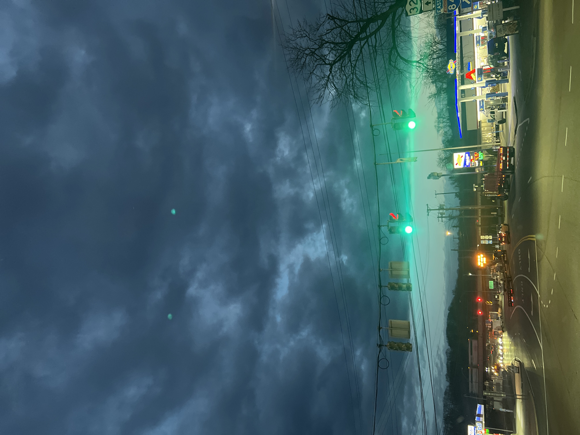 The image shows a street scene at dusk. On the right side of the image is a road with traffic. A Sunoco gas station is brightly illuminated, with fuel prices visible on a sign. On the left, the sky is dark and cloudy. Traffic lights are visible along the road, some displaying green and red signals. Power lines stretch across the scene. The location may be the City of Newburgh, United States of America.