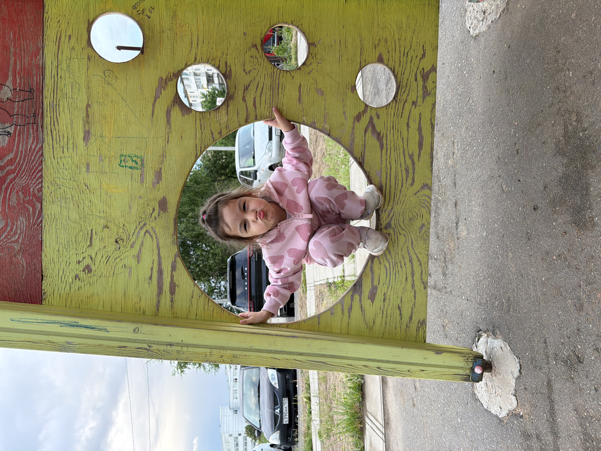 The image captures a young child peeking playfully through a large circular cutout in a weathered, lime-green wooden structure. The child, dressed in a pink jumpsuit with white heart patterns and white sneakers, is positioned horizontally in the frame, making a "kiss" face with pursed lips while holding onto the edges of the opening.
The wooden structure, exhibiting peeling paint and visible wood grain, features several smaller circular cutouts above the child, which reflect glimpses of the bright, possibly overcast, sky, distant apartment buildings, and parked vehicles. Indistinct scribbles and markings, including what resembles "117" and other graffiti, are present on the green surface. A red wooden panel is partially visible on the left side of the structure.
The scene is an outdoor setting, likely a public space or playground, adjacent to an asphalt ground surface. In the background, visible through the cutouts and overall scene, are a white van and a black car. The black car's license plate displays the text "E390AK 159," with "159" indicating the Perm Krai region of Russia, consistent with the location of Chaikovsky. The time of day is daylight.