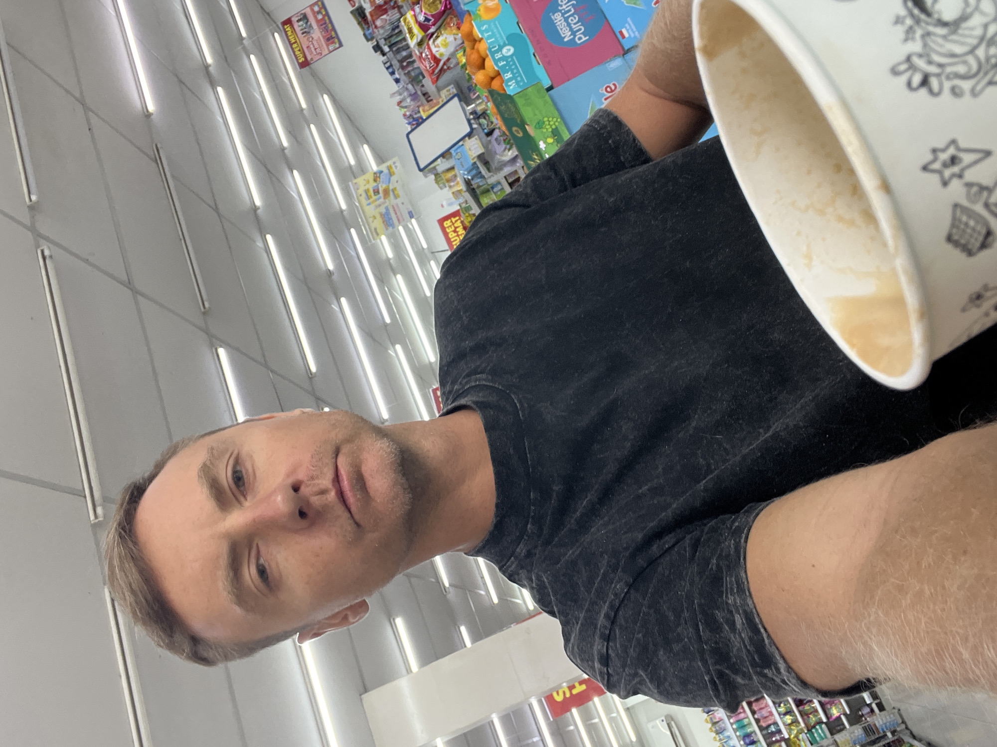A man with short, blond hair and a slight stubble is taking a selfie in what appears to be a grocery store. He is wearing a dark, t-shirt with a textured pattern. In the foreground, his right arm is visible, holding a white paper cup with a decorative pattern. The background shows shelves stocked with various products, overhead fluorescent lighting, and a sign that reads "SUPER HEMAT". The overall scene suggests a casual shopping trip in a commercial establishment in Sukawati, Indonesia.
