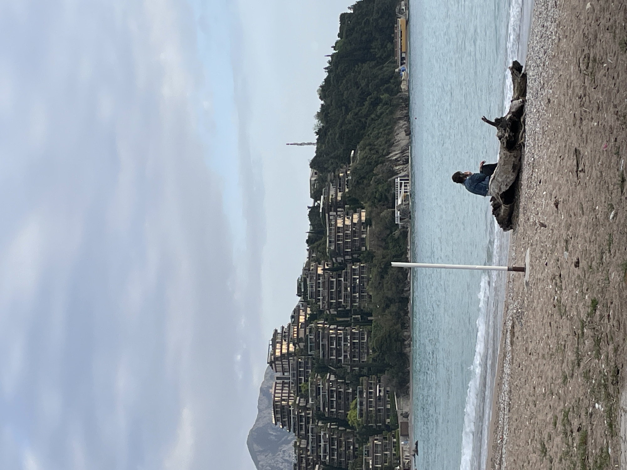 A beach scene in Budva, Montenegro. The buildings are modern structures built on a steep hillside. A person is sitting on a piece of driftwood on the beach. There are mountains in the background. The weather is overcast. There is a pole on the beach near the waterline.
