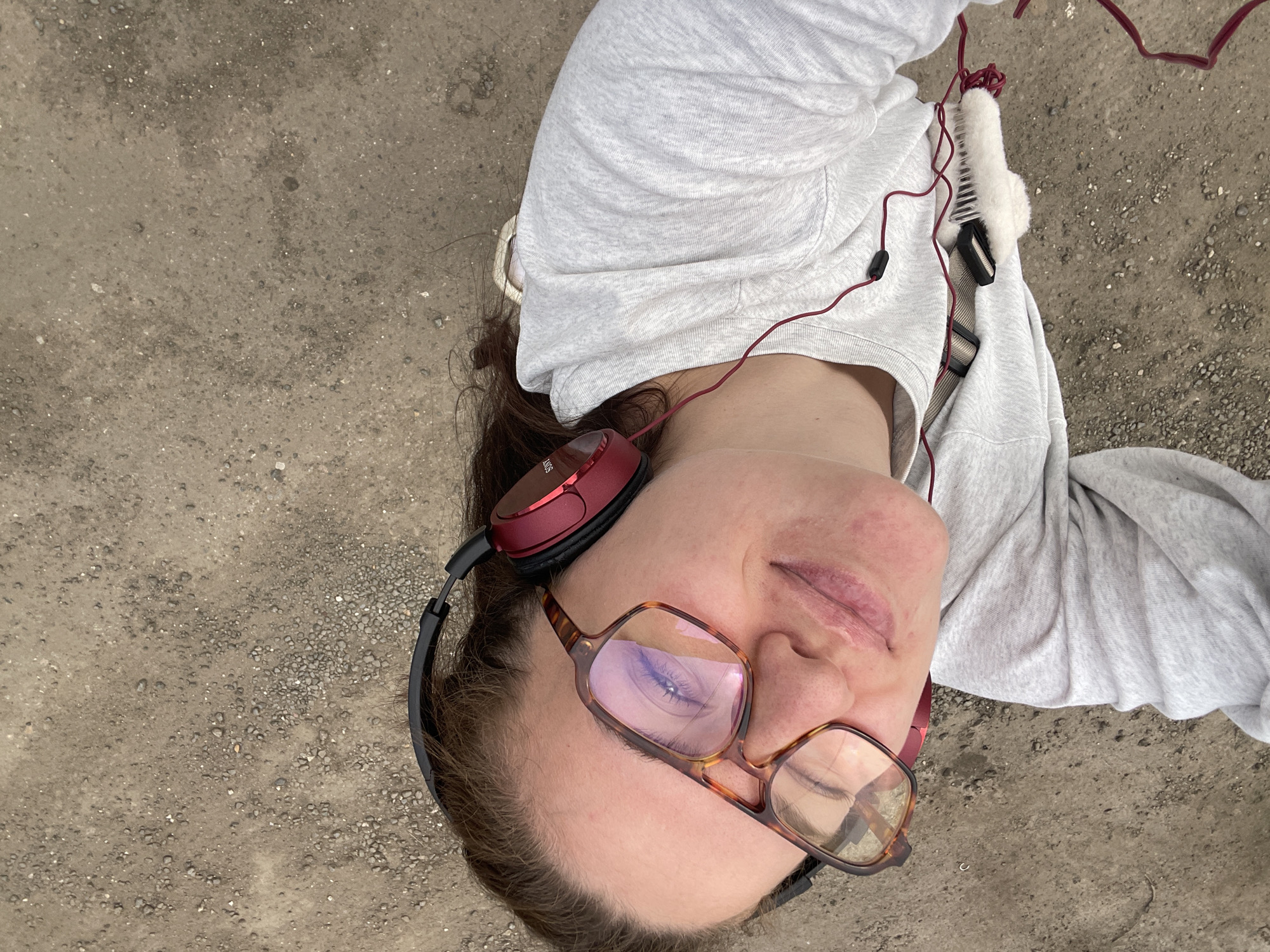 The image shows a person lying on a gray, textured surface, likely concrete or asphalt. The person is wearing tortoiseshell glasses, red headphones, and a light gray hoodie. Red wired headphones are connected to a device or source out of view. A white fluffy object with a black clip is attached to the hoodie. The person's head is tilted towards the camera. The location is not discernible as Molokovo, Russia, as there is no contextual information to confirm this location.