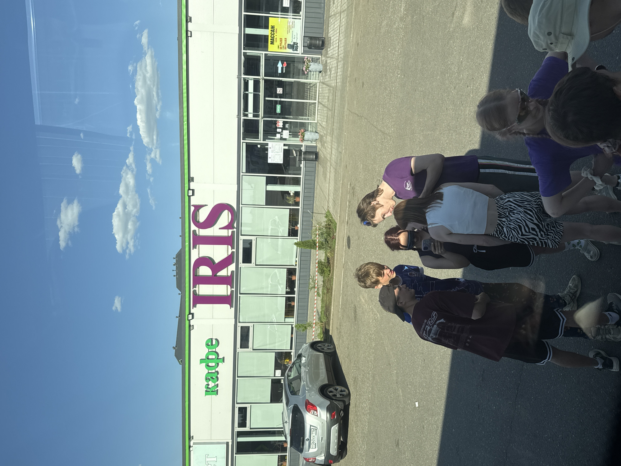 The image shows a group of people standing on a paved surface next to a building. The building has the word "IRIS" written vertically in large letters, with the word "KAFE" (cafe) below it in smaller letters. The building appears to be a business, possibly a cafe or store. A silver car is parked in front of the building. The sky is visible in the background, and there are some clouds. The image was likely taken in Казань, Russia, based on the Cyrillic lettering of the license plate of the car. The group of people seems to be looking at something or someone in front of them.