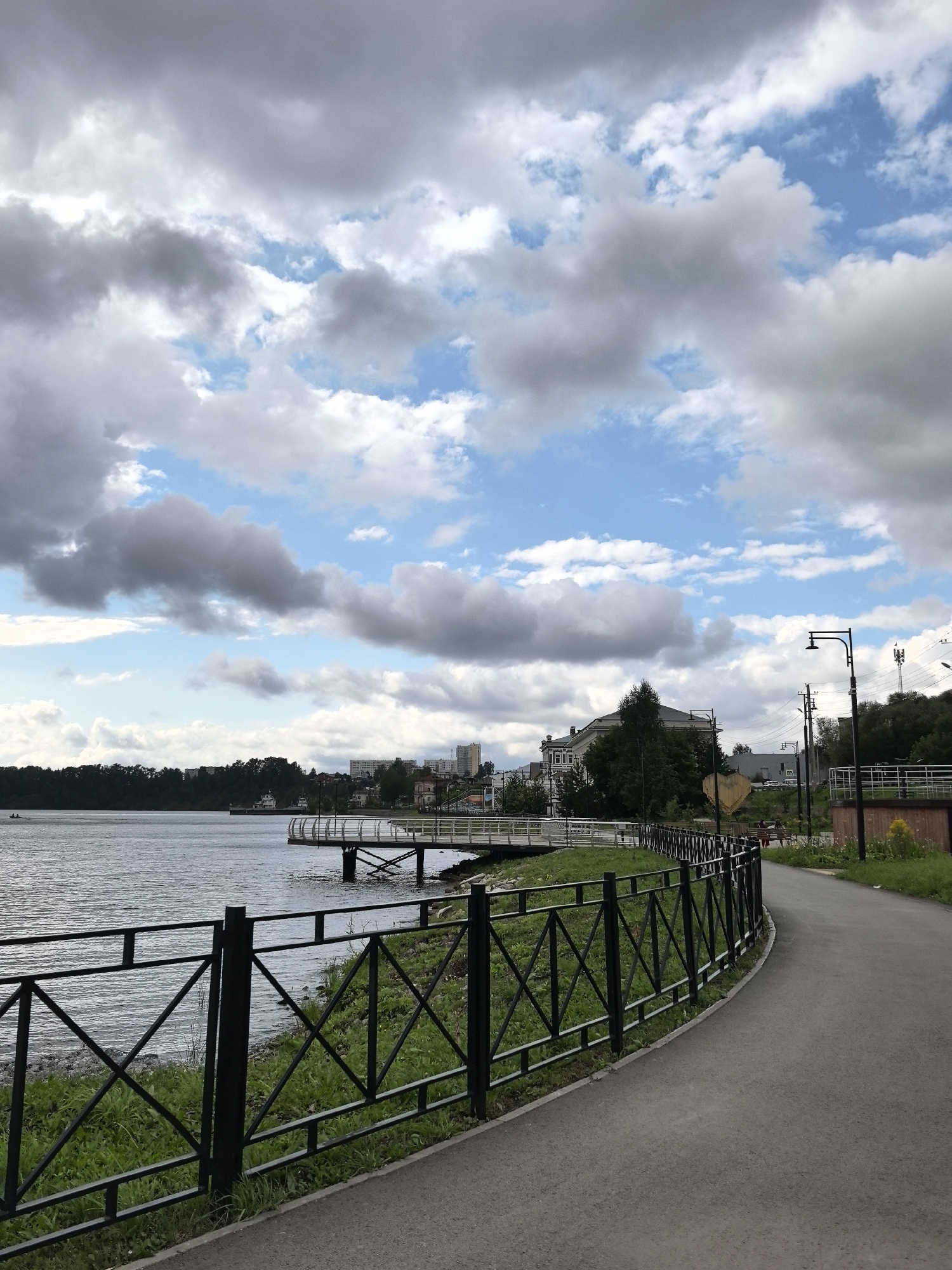 This image captures a view of the Kama River embankment in Dobryanka, Russia, on a cloudy day. A paved walkway follows the curve of the river, bordered by a black metal fence. Lush green grass grows behind the fence, and a body of water stretches out to the left. In the distance, across the river, a line of trees forms the horizon, with buildings and structures visible along the shoreline. A white pier extends into the water, and a small boat can be seen in the distance. Several lampposts line the walkway. The sky is filled with a mix of gray and white clouds, interspersed with patches of blue.