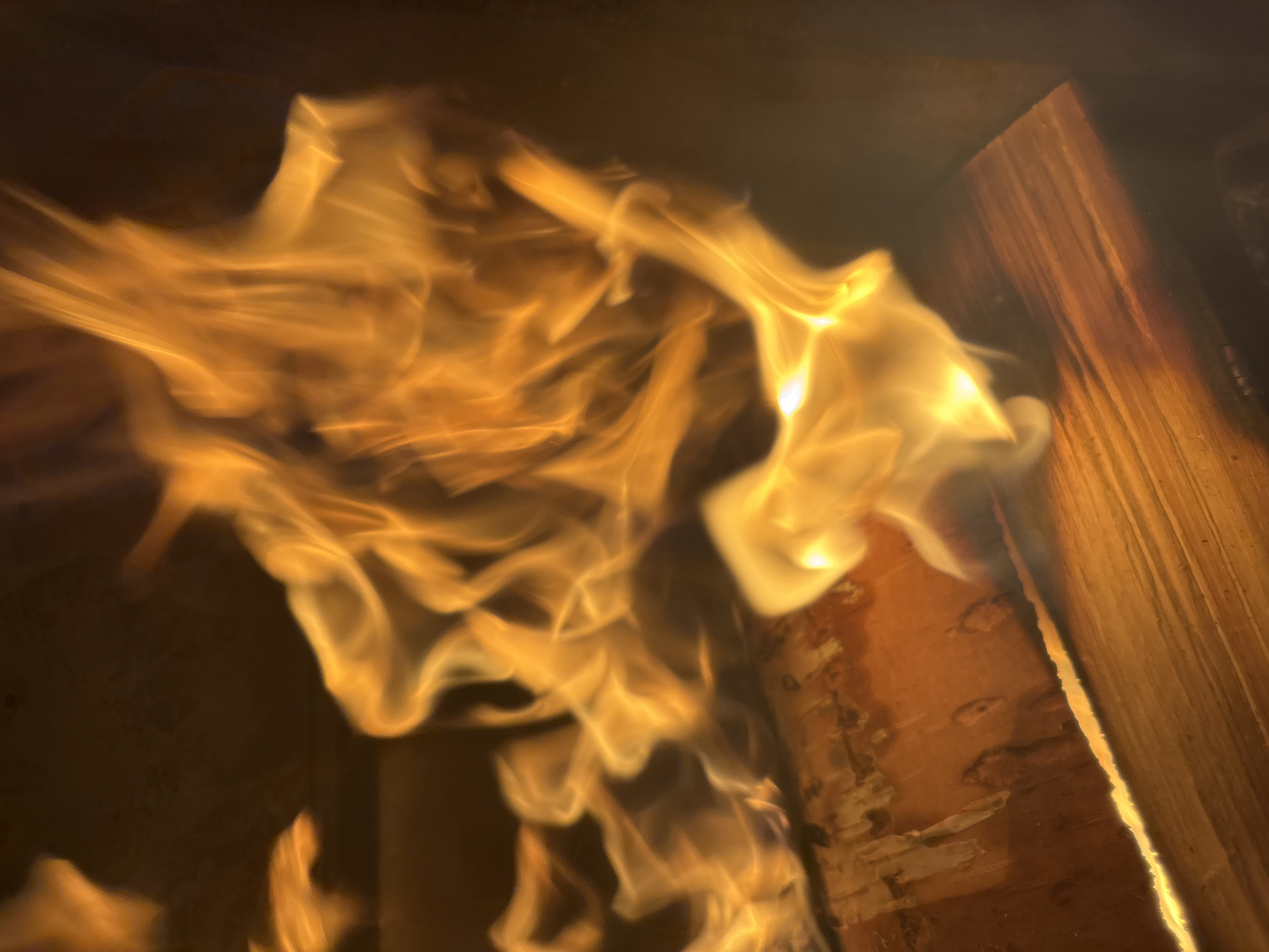The image shows a close-up view of a fire burning on wood. The flames are bright orange and yellow, with tendrils reaching upwards. The wood appears to be logs or planks, with visible grain and texture. The background is dark and out of focus, suggesting an indoor setting or a dimly lit area. There are no people visible in the image, nor any indication of a specific location within the city of Zvenigorod, Russia. The scene is solely focused on the fire and the burning wood.