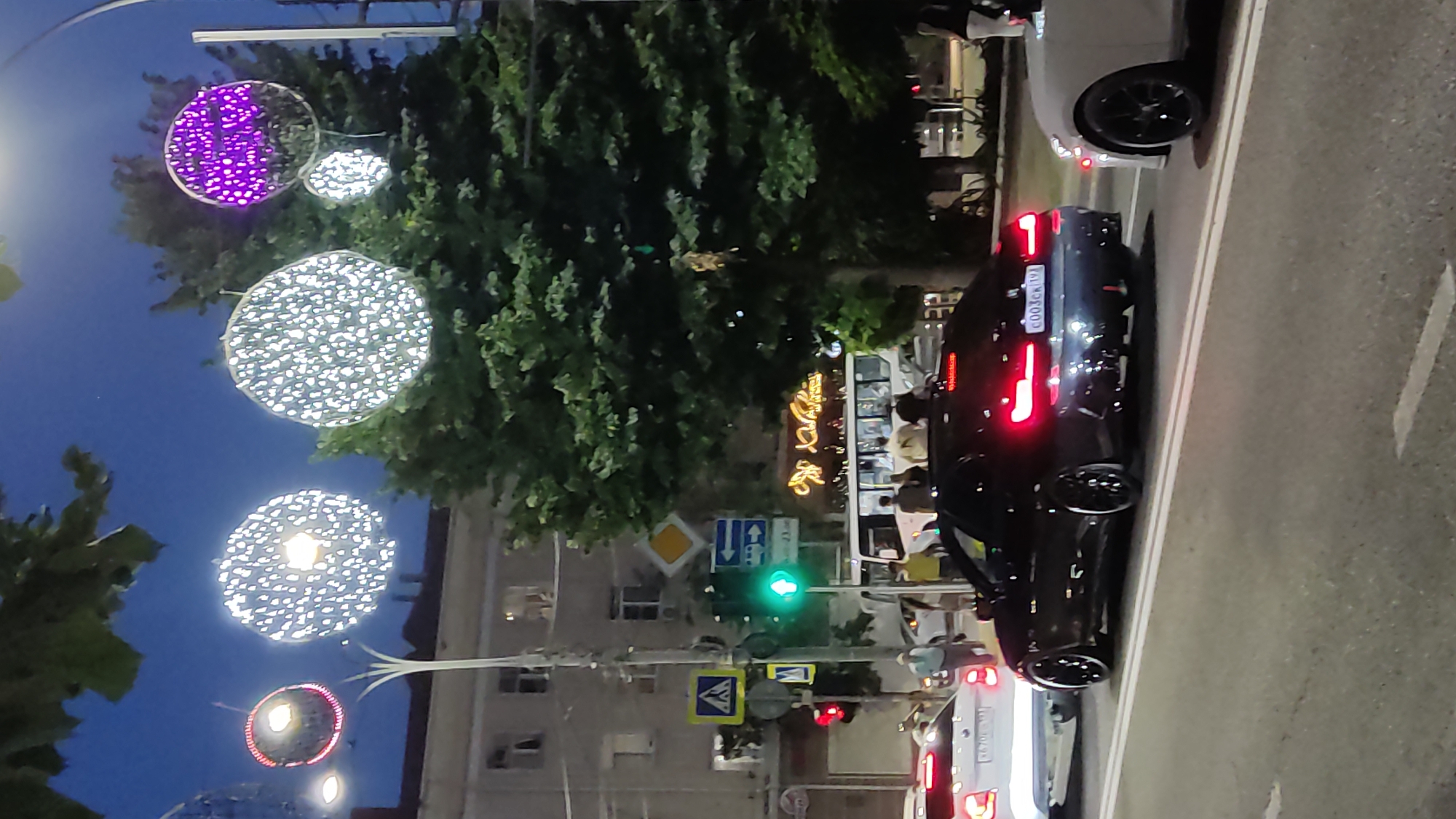 The image depicts a street scene at dusk in Krasnodar, Russia. Ornate circular lights, some illuminated with white and purple LEDs, are strung across the street, adding a festive ambiance. Several vehicles, including a black BMW coupe with illuminated taillights and a white sedan, are on the road. Pedestrians are visible on the sidewalk near a bus. Traffic signals and street signs are present, indicating a busy intersection. A large tree with dense foliage stands to the right of the frame. Buildings are visible in the background.