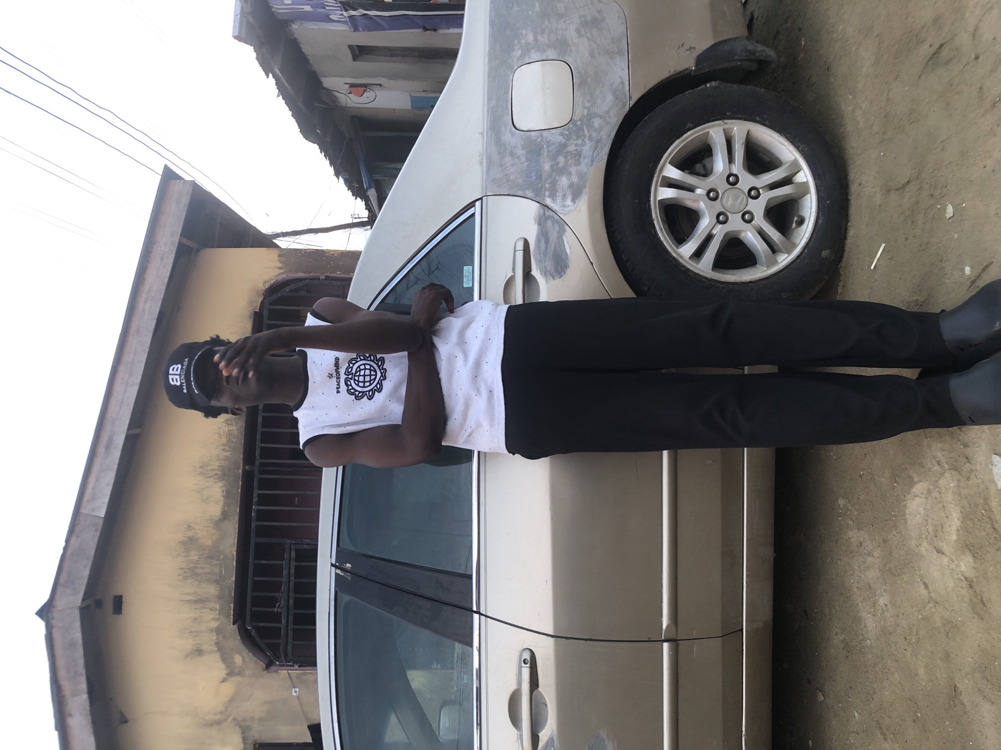 A young man stands in front of a silver sedan, parked on a dirt road in Ado, Nigeria. He wears a black baseball cap with a white logo and a white sleeveless t-shirt with a graphic design. His arms are crossed, and he is holding a small object in his right hand, near his face. The car has a slightly damaged exterior, with visible repair marks on the rear quarter panel. Behind the car, a building with yellowed walls and a corrugated metal roof is visible, along with other structures in the background that suggest a residential or commercial area. The sky is overcast, and the overall lighting suggests it is daytime.