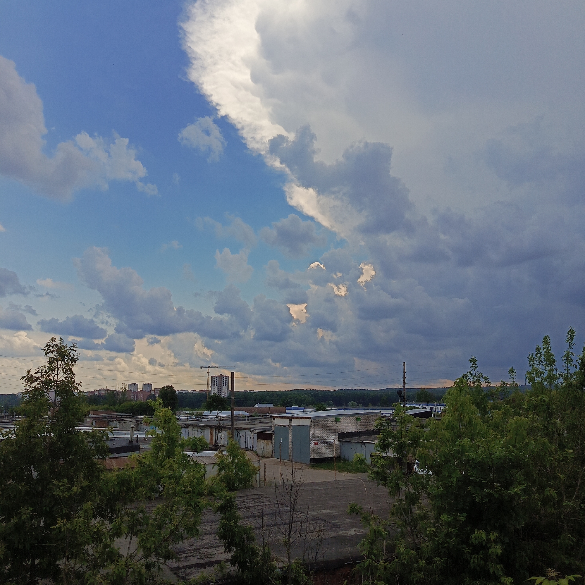 This is a medium-distance, high-angle, full shot of a collection of garages and buildings in Chaikovsky, Russia, under a dramatic, cloudy sky. The sky occupies the upper two-thirds of the frame, dominated by a large, bright white cloud formation on the right, with a transition to darker, more ominous grey clouds extending across the top and upper left. The lower third of the image shows a cluster of flat-roofed, rectangular garages, many with metal roll-up doors. Some garages appear to be made of concrete blocks, while others have corrugated metal siding. In the background, a line of trees separates the garage area from a distant cityscape with several multi-story buildings and a construction crane. The foreground is partially obscured by the dense foliage of green trees and bushes, casting shadows on the ground. The ground surface in front of the garages appears to be asphalt or concrete, with some visible cracks and debris. No people or animals are visible in the scene. The lighting suggests it is either early morning or late afternoon, with the sun breaking through the clouds in some areas.