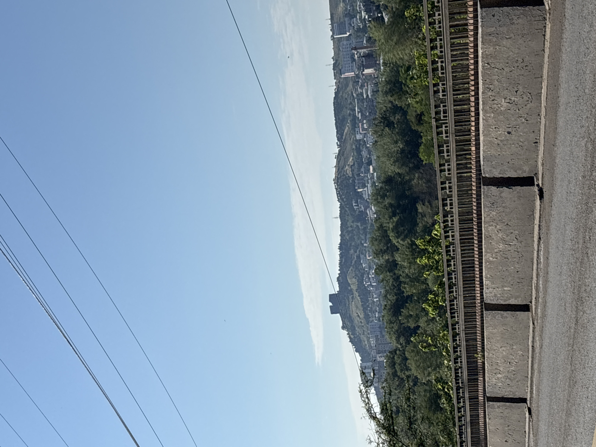 A wide shot captures a cityscape atop a hill in Tbilisi, Georgia, under a clear, pale blue sky. In the foreground, a grey concrete barrier runs horizontally, with a black asphalt road in front of it. Above the barrier, a decorative metal fence lines a bridge. Behind the fence, a dense line of green trees forms a vibrant backdrop. The middle ground features a city with buildings clustered on a hilly terrain. Dominating the crest of the hill are the ruins of a stone fortress, with two prominent square towers. The sky is mostly clear, with a few wispy clouds and several utility wires stretching across the frame.