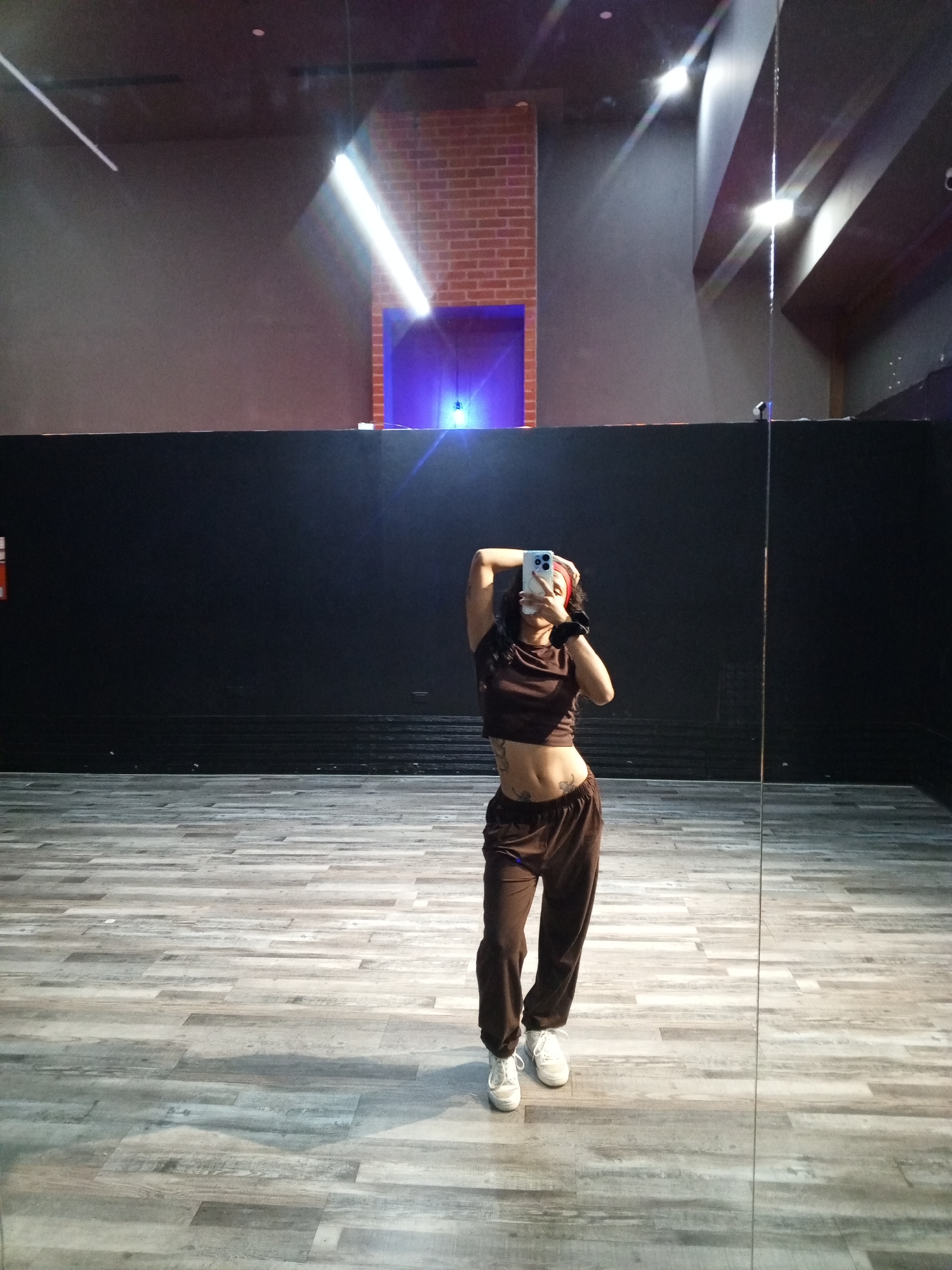 A woman stands in a dance studio, taking a selfie in a mirror. She is wearing a brown crop top and brown sweatpants, with white sneakers. She has a red headband and her hair is pulled back with a scrunchie. The studio has a gray wood-look floor, a black wall, and a brick feature with blue lighting. The ceiling has exposed beams and track lighting. The location is Pampatar, Venezuela.