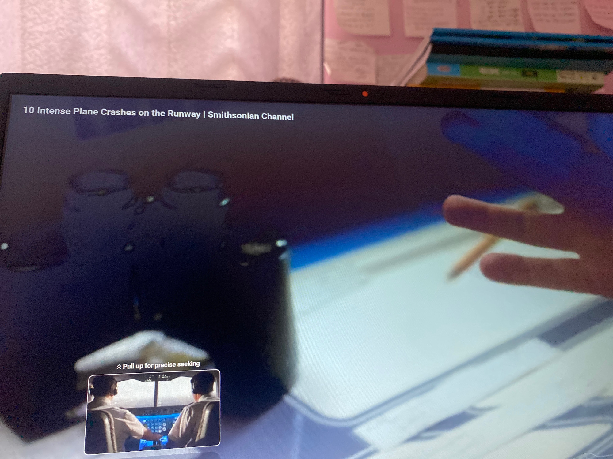 The image shows a laptop screen displaying a video from the Smithsonian Channel titled "10 Intense Plane Crashes on the Runway". The video appears to be a simulation or documentary showing the interior of an aircraft cockpit with two pilots. In the foreground of the video, a small inset window shows the pilots, one in the captain's seat and the other in the co-pilot's seat, wearing white shirts and headsets. They are looking forward, presumably at the runway. A text overlay on the inset reads "Pull up for precise seeking".
The main part of the video displays a blurred, dark image that seems to be a reflection in the cockpit window, with elements of the surrounding environment visible. There is also a close-up of a hand holding a pencil and interacting with a tablet or screen in the upper right portion of the laptop display.
The background of the overall image shows the top of the laptop and a shelf with a stack of books. The laptop has a small red indicator light, likely a webcam light, near the top bezel. The environment suggests an indoor setting, possibly a room where the laptop is being used. There is no information within the image that indicates a location in Hemayetpur, Bangladesh.