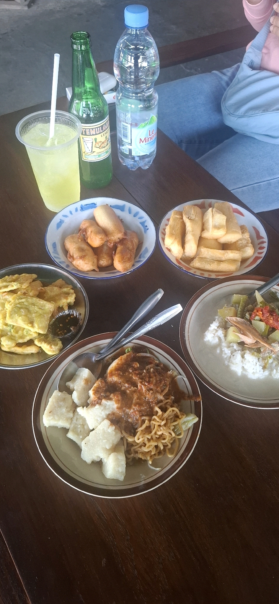 In Desa Sobokerto, Indonesia, a wooden table is laden with food and drinks. A clear plastic cup filled with a yellow, iced beverage and a white straw sits next to a green glass bottle of Temulawak Lemon drink. A clear plastic bottle of Le Minerale water is also present. In front of the drinks, several plates of food are arranged. One plate holds pieces of fried banana, another contains several pieces of fried cassava. A metal dish contains fried tempeh with a small dish of dipping sauce. A round plate in the foreground features a portion of rice topped with a savory sauce and noodles, accompanied by several pieces of white, cubed food, likely lontong or rice cake. Another round plate to the right holds white rice mixed with vegetables and a piece of dried fish. Two spoons and two forks rest on the table, indicating a meal for at least two people. The background shows a person in blue jeans and a pink top, with a blue bag slung over their shoulder. The setting appears to be an outdoor or semi-outdoor dining area, with a wooden bench visible behind the table.