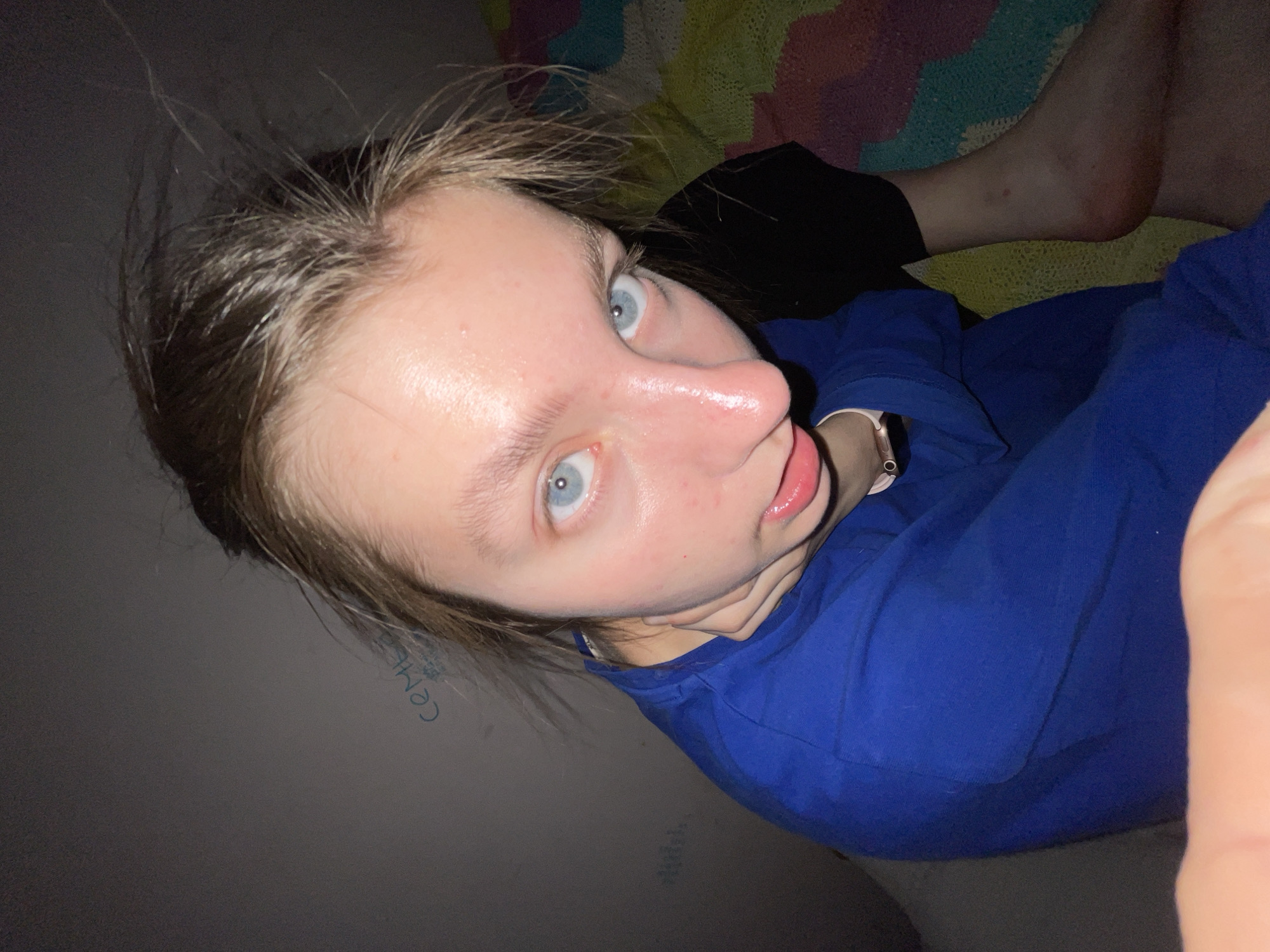 A pale-skinned young person with blue eyes and light brown hair is positioned at an angle in the frame, appearing to be taking a selfie. The person is wearing a blue shirt and a black garment, possibly shorts, is visible. A limb of another person is visible in the background, lying on top of a patterned blanket. The background also includes a wall with faint writing that appears to say "CEM6E." The image is taken in an indoor environment. There is no indication of the location being in the city of Moscow, Russia.