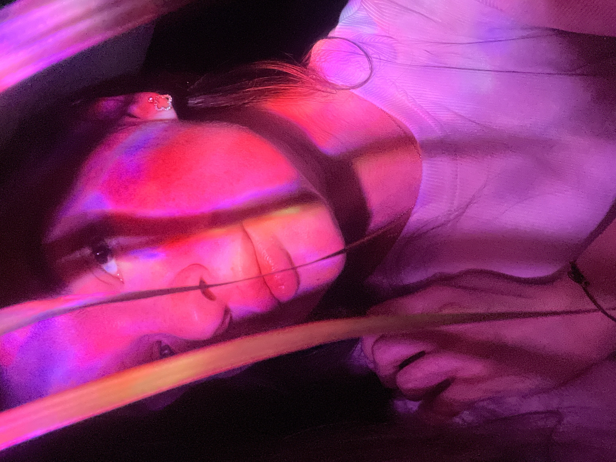 A person lies on their back with their head at the top left of the frame, illuminated by a red and purple light. Part of their face is obscured by what appear to be long, thin leaves. There is a hair clip with a bejeweled design in their hair. The person wears a light-colored shirt or dress. The background is predominantly dark. It's not possible to determine the city of Kazan, Russia, from this image alone.