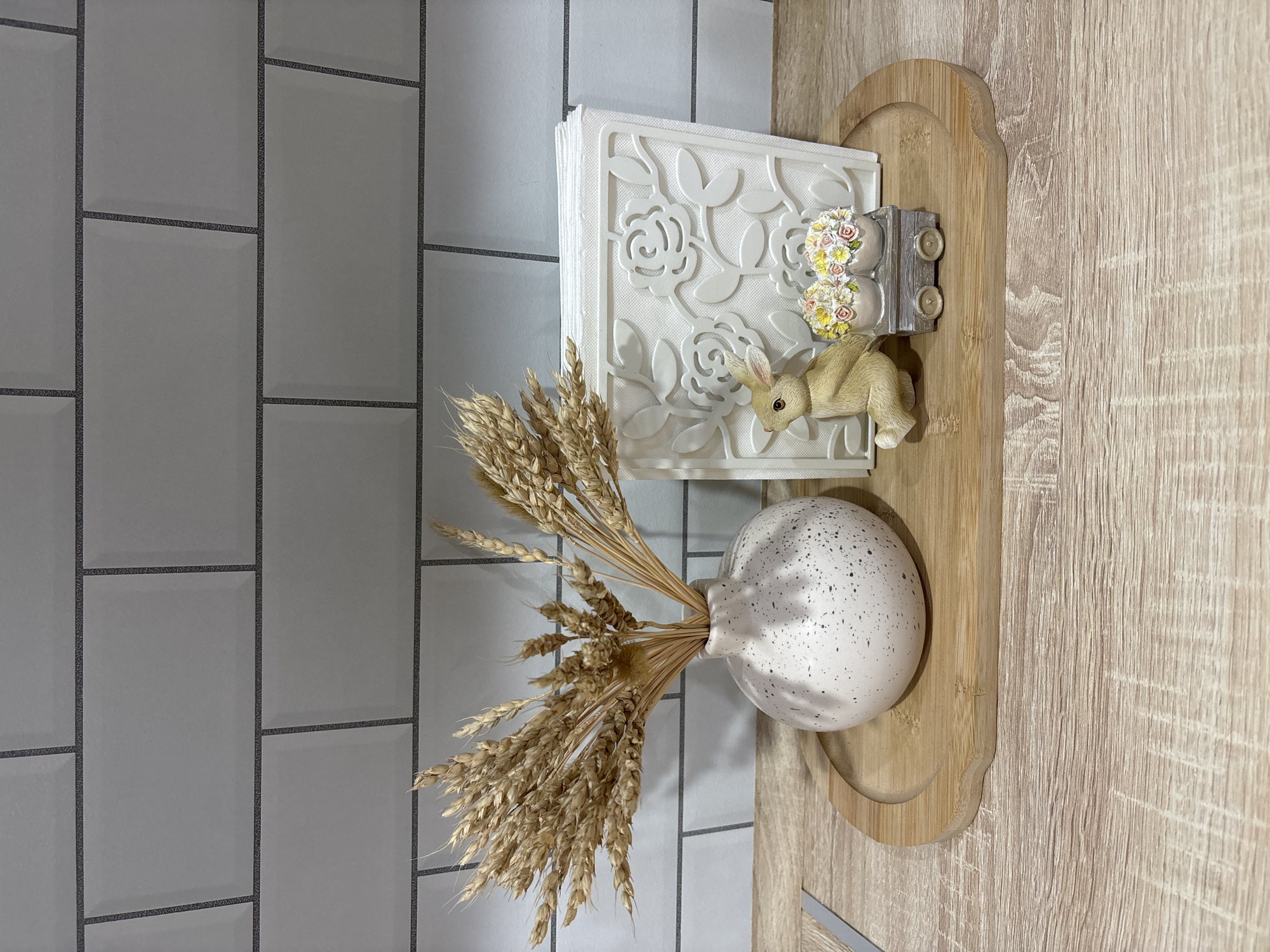 The image shows a still life arrangement on what appears to be a light wood-grained surface. From left to right, there is a background of gray subway tile. A vase filled with wheat stalks rests on the wooden surface. A stack of white napkins within a decorative white holder is placed near the vase. A small decorative bunny figurine is on top of the napkins, next to a decorative miniature car with floral accents. The objects are situated in a way that suggests a staged, possibly decorative, scene. There is no indication of the location of Noviy, Russia, within the image.