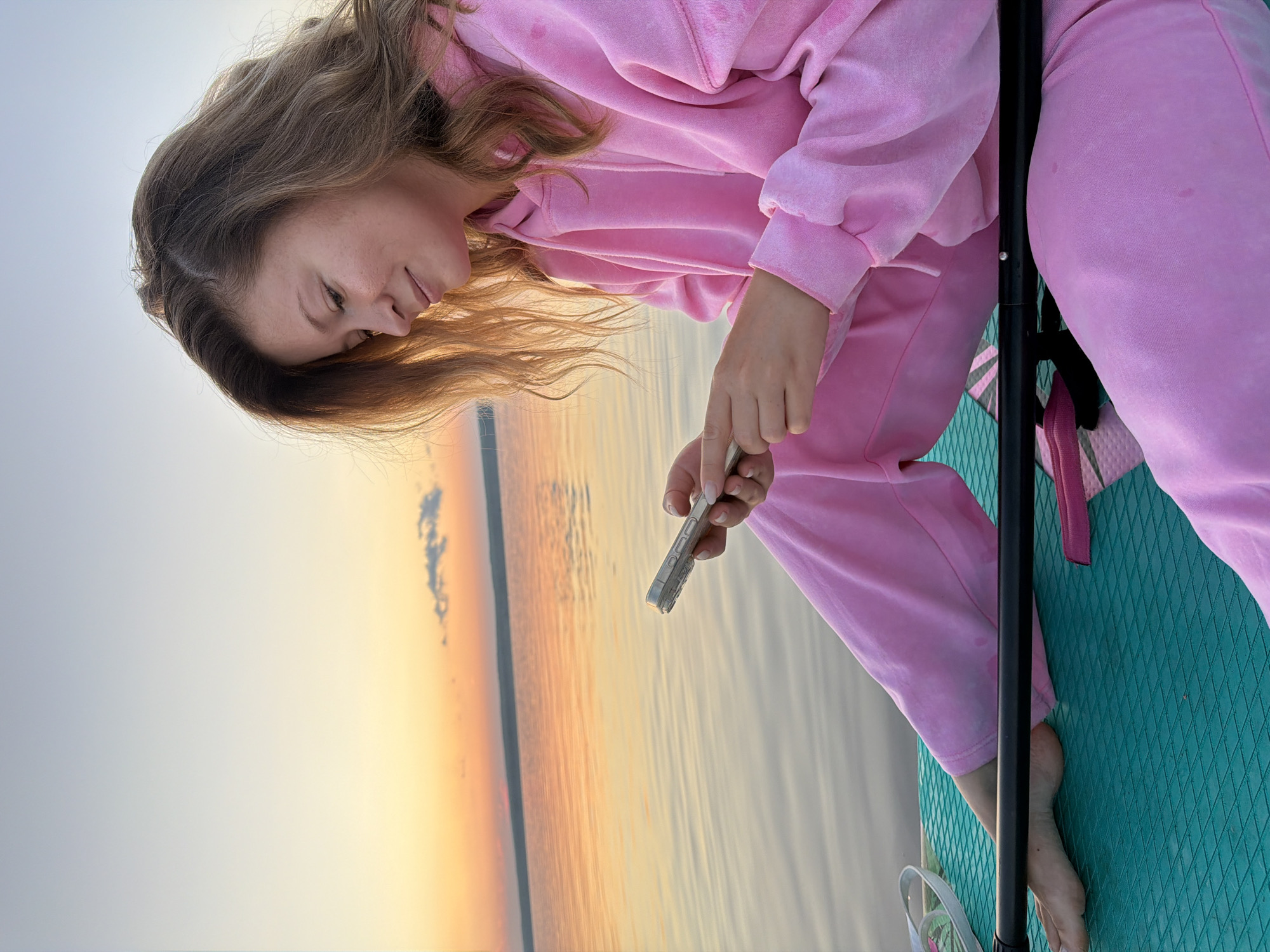 A young woman with light brown hair is sitting on a teal paddleboard in the water at sunset, looking down at her phone. She is wearing a pink velour hoodie and pants. The sky is a gradient of pale blue to orange, with a faint cloud on the horizon. The water is calm and reflects the colors of the sky. A black paddle lies across her lap. The scene appears to be a tranquil moment, possibly during a paddleboarding excursion on the Kama River in Chaikovsky, Russia.