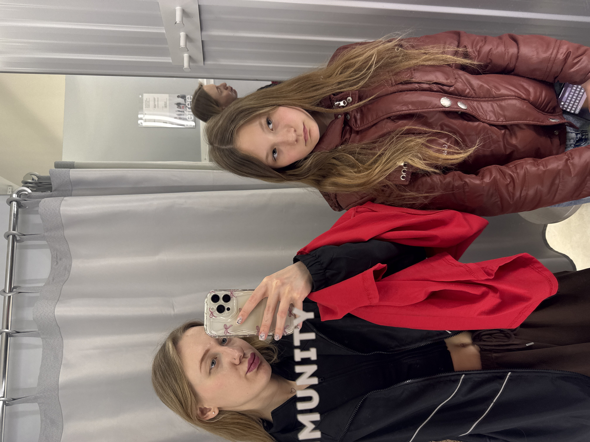 A woman and a young girl are in a dressing room, likely in a retail clothing store. The woman, positioned lower in the frame, holds a smartphone with a clear case decorated with bows, taking a selfie in the mirror. She wears a black jacket with white stripes, a black top with "MUNITY" printed on it, and has a red garment draped over her shoulder. The young girl, positioned above her in the frame, wears a maroon puffer jacket. A gray curtain is visible on the left. The location appears to be indoors, within the city of Rostov-on-Don, Russia.
