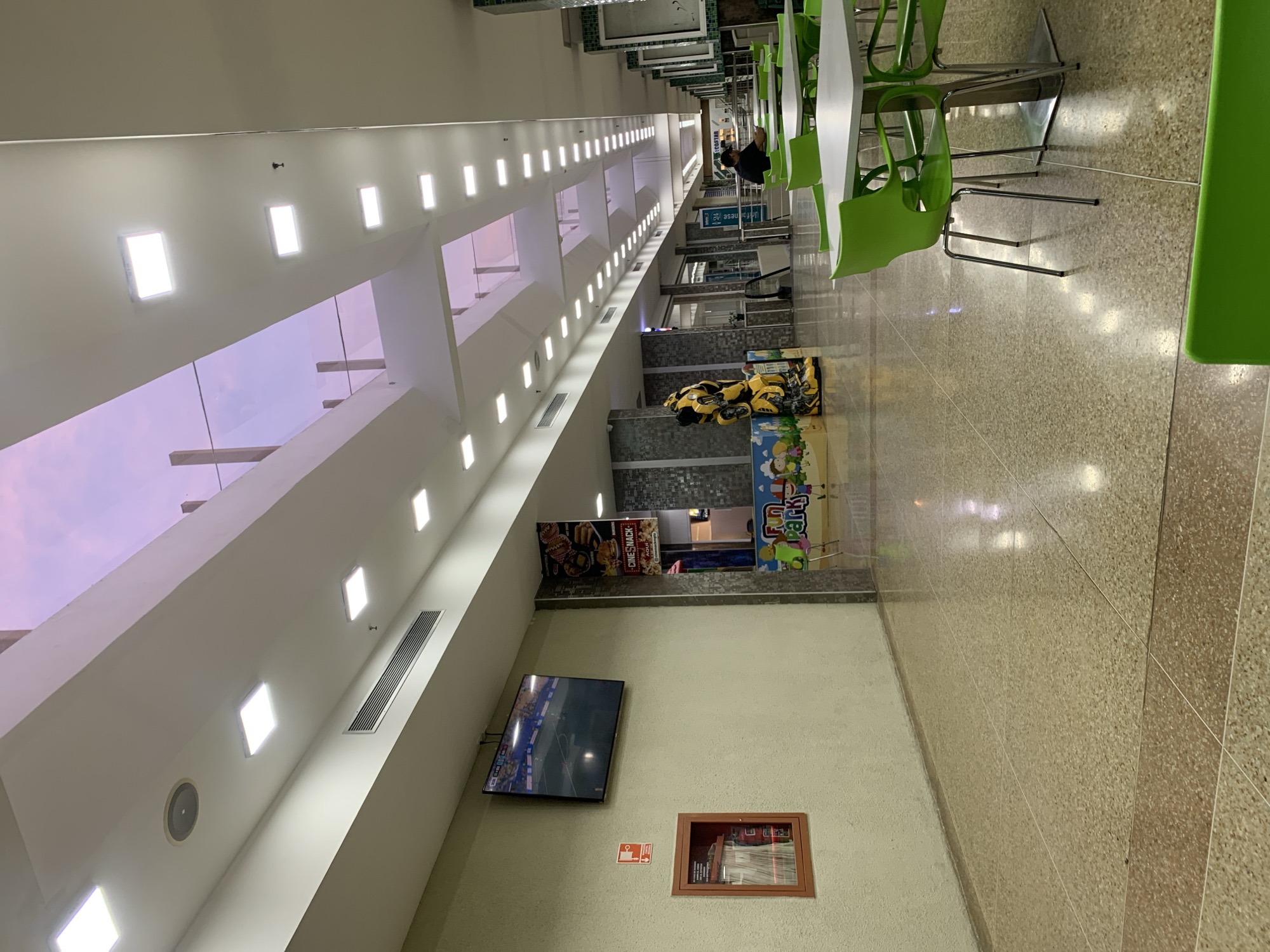 The image depicts a section of an indoor commercial space, likely a mall or food court, with a view towards a skylight. The ceiling features numerous recessed square lights and air vents. A large television screen is mounted on the left wall, displaying a sports match. Below the TV is a red fire extinguisher cabinet. Further down the wall, a colorful mural depicting cartoon characters and a yellow transformer statue are visible.
The floor is paved with speckled terrazzo tiles, with a darker band of tiles running across it. To the right, several tables with bright green chairs are arranged, some occupied by patrons. The environment appears well-lit, with natural light filtering through the skylight, revealing a purple and pink sky. The overall impression is one of a casual, public space, possibly a recreational or dining area within a larger establishment. The provided context places this scene in El Valle del Espíritu Santo, Venezuela.