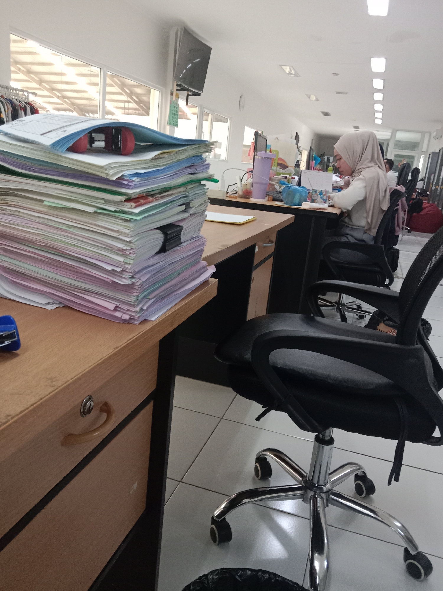 The image depicts an indoor scene that appears to be an office environment in Semarang, Indonesia. In the foreground, a large stack of papers sits on a wooden desk. The papers are of various colors, including white, pink, blue, and green, and are bound together with a black clip. A red object, possibly a stapler, is visible on top of the stack. A blue stapler is also present on the desk to the left of the paper stack.
In the midground, a woman wearing a light brown hijab is seated at another desk, facing away from the camera. She is looking down at papers on her desk and appears to be working. On her desk, there are various office supplies including a purple water bottle, a calendar, and some pens. To her right, a black office chair with a chrome base and wheels is prominently featured, facing towards her desk. Another similar chair is visible behind it.
The background shows a long office space with multiple desks and chairs, illuminated by fluorescent ceiling lights. A television screen is mounted on the wall in the upper left portion of the image. Rows of clothing racks with garments are visible near a large window in the upper left. The floor is tiled with white square tiles. The overall impression is that of a busy administrative workspace.
