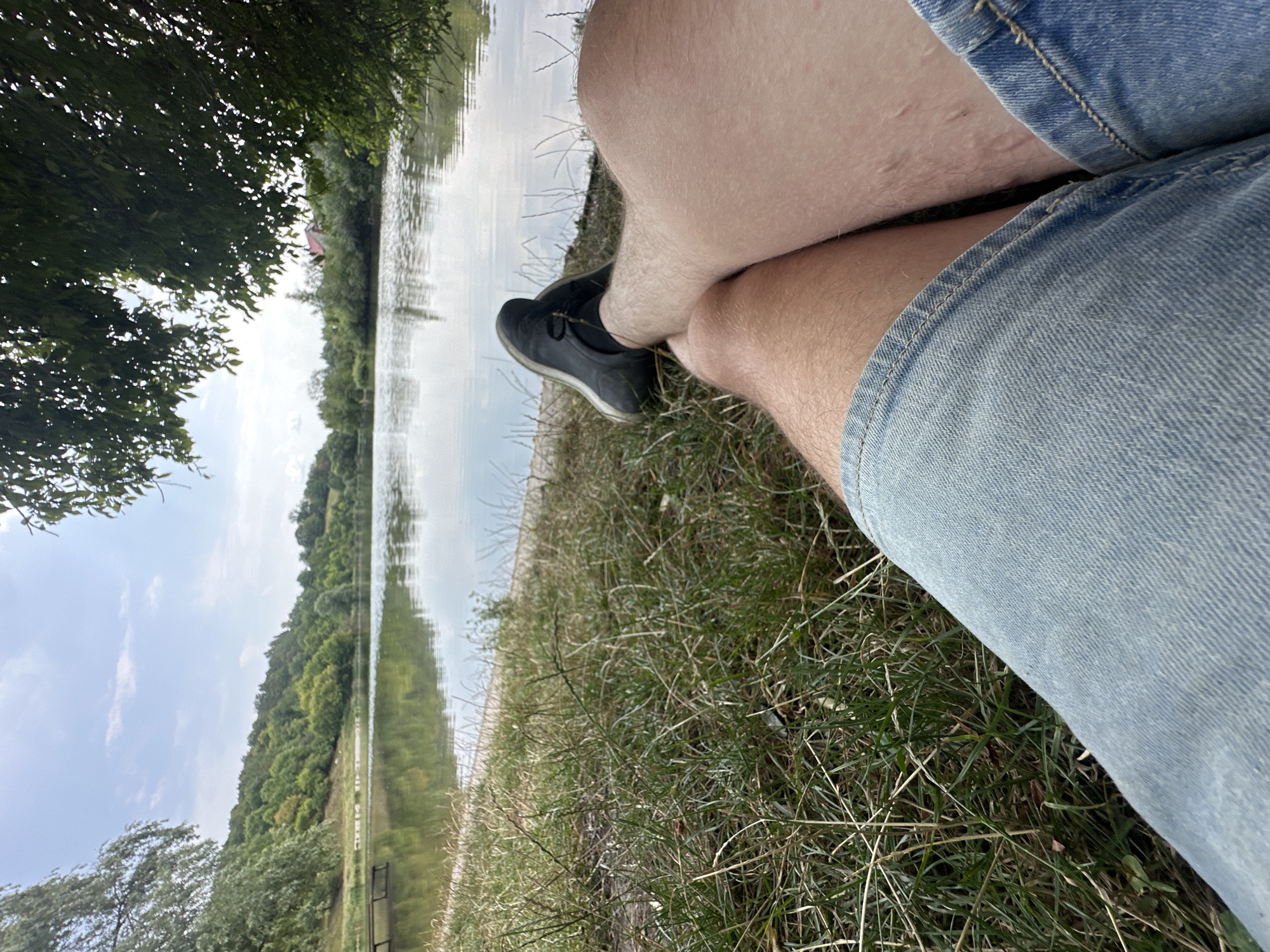 The image depicts a personal perspective of a tranquil lakeside scene. The foreground is dominated by the legs of a person, clad in denim shorts and black shoes, resting on a grassy bank. The person's legs are slightly tanned and hairy. The grass appears dry and somewhat sparse, with scattered blades of straw-like vegetation.
Beyond the immediate foreground, the calm surface of a lake reflects the overcast sky and the lush, tree-covered hills that form the backdrop. On the left side of the lake, a wooden dock is partially visible, jutting out into the water. The trees on the hills display various shades of green, suggesting a summer or early autumn setting. A few scattered buildings can be seen nestled among the trees on the far side of the lake. The overall impression is one of peace and natural beauty. The location is identified as Makiv, Ukraine.