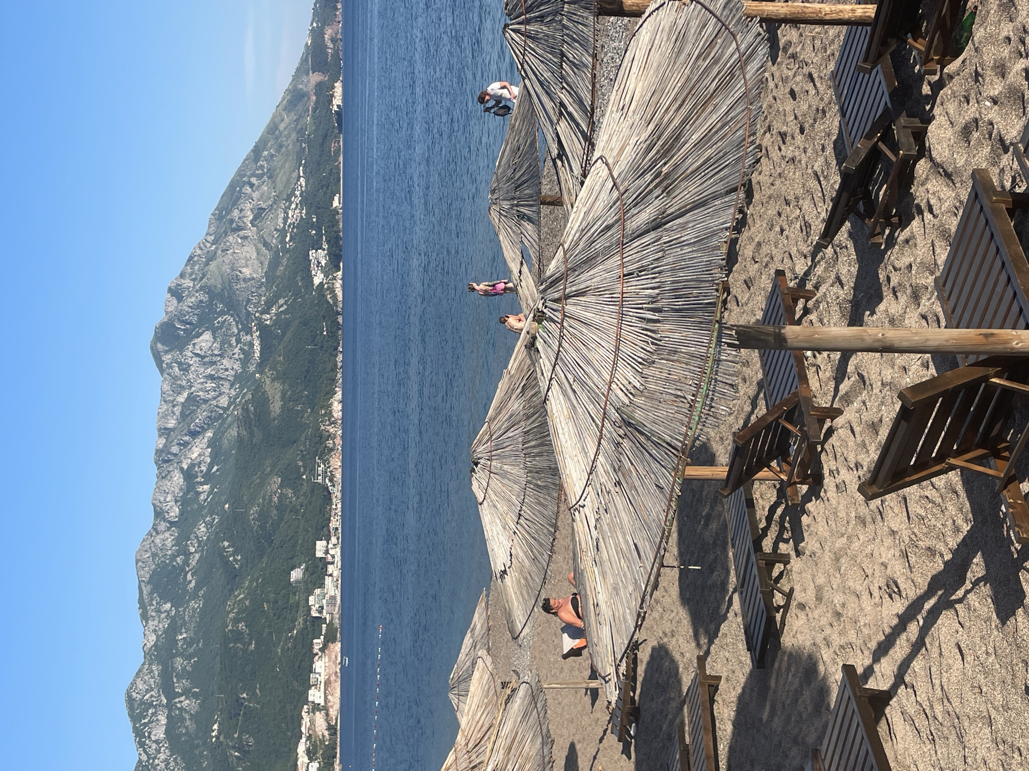 Captured from an elevated perspective, the image shows a beach scene in Boreti, Montenegro. Straw umbrellas provide shade over lounge chairs on a sandy beach. People are on the beach under the umbrellas. In the background, the Adriatic Sea extends to the base of a large, mountainous terrain where buildings are visible. The sky is clear and blue.