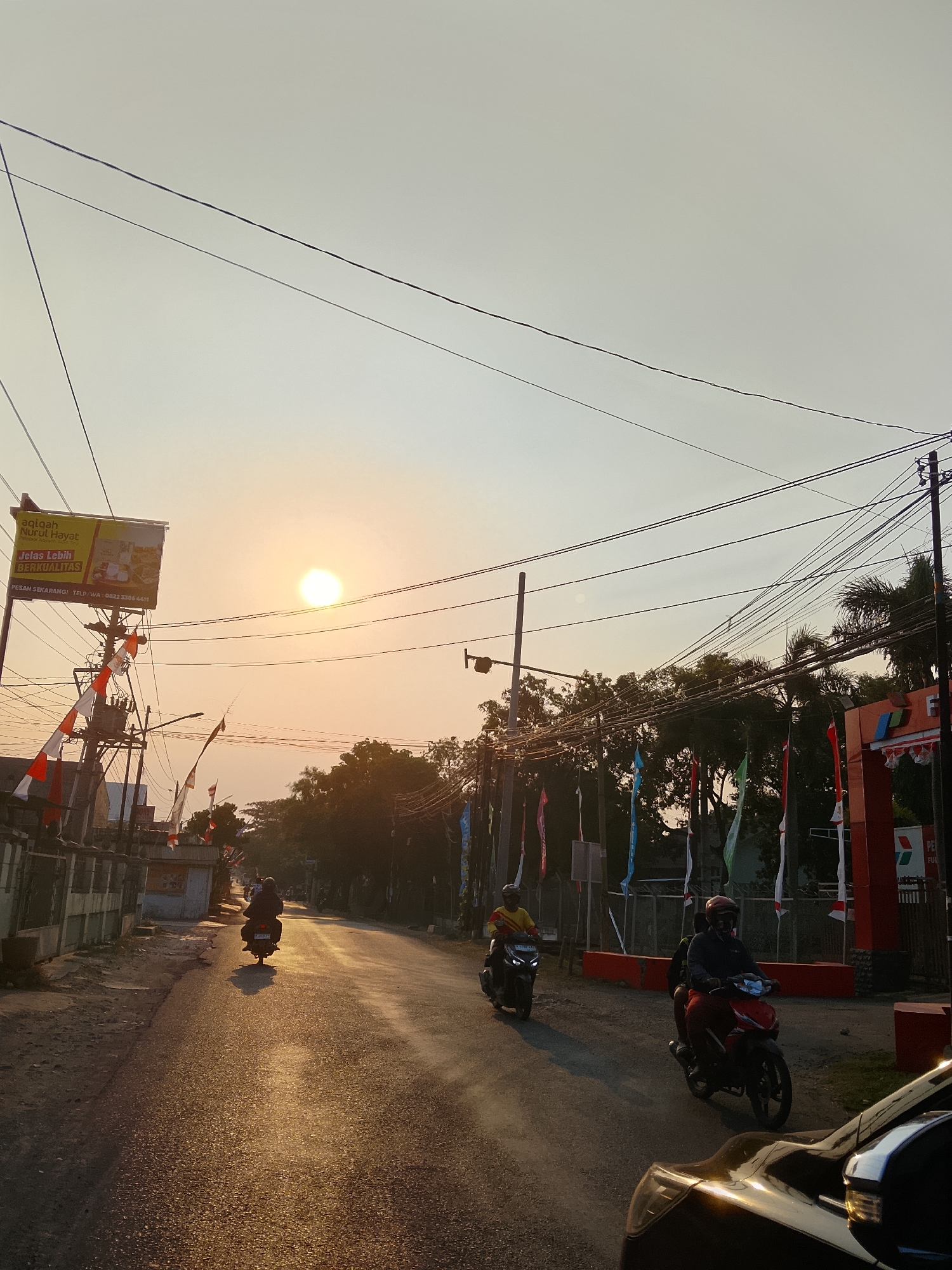 The image depicts a street scene at sunset in Ngelo, Indonesia. The sun is low in the sky, casting a warm, golden light on the asphalt road and the surrounding environment. Several Indonesian flags, red and white, are visible along the roadside, attached to poles and fluttering in the breeze. Power lines crisscross the sky, casting shadows.
In the foreground, a black car is partially visible on the right side. On the road, three motorcycles are in motion. The closest motorcycle, on the right, carries two people, the driver wearing a dark jacket and red pants, and the passenger behind them also wearing dark clothing. Further down the road, another motorcycle with a single rider wearing a yellow shirt and a helmet is approaching. In the distance, a third motorcycle with a single rider is heading away from the camera.
To the left of the road, there is a building with a large advertisement banner. The banner, primarily yellow, has text in Indonesian, including "aqiqah Nurul Hayat" and "JELAS LEBIH BERKUALITAS". A power transformer and electrical equipment are mounted on a utility pole near the banner.
On the right side of the road, a red structure, possibly an entrance arch, with a "Pertamina" logo is visible. Trees and foliage line the sides of the road, with some appearing to be palm trees. The overall atmosphere is that of a typical street in a Indonesian town during a beautiful sunset.
