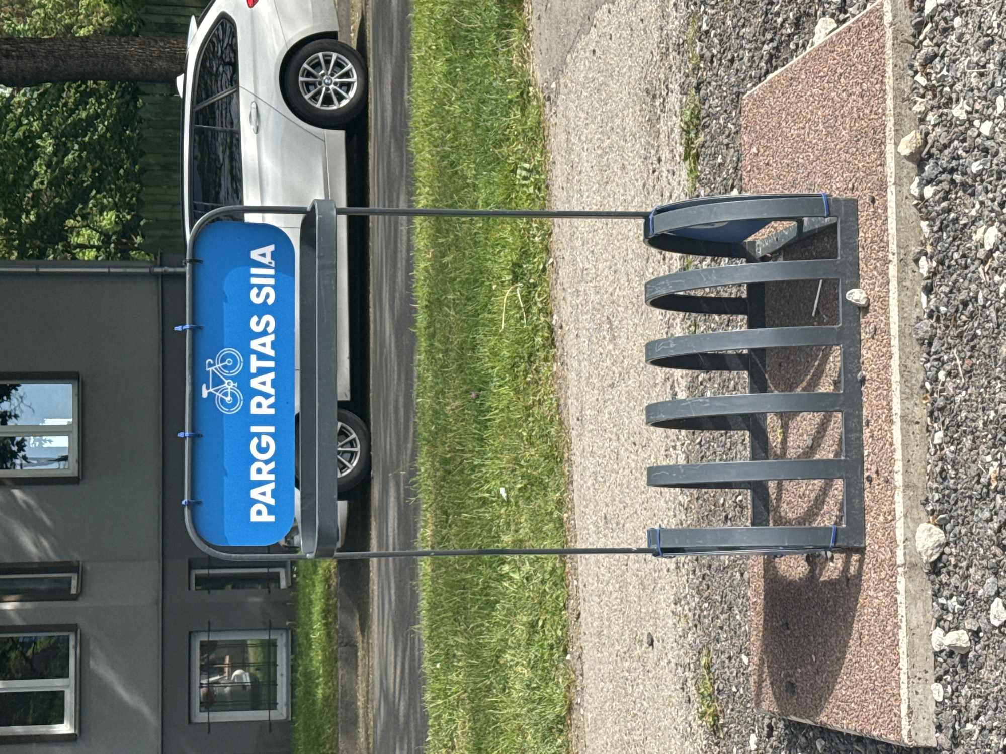 The image depicts an outdoor scene in Tallinn, Estonia, featuring a bicycle rack with a blue sign that reads "PARGI RATAS SIIA". The bicycle rack is made of dark metal and has space for several bicycles. Behind the rack, a white car is parked on a paved surface. The background includes a building, a patch of grass, and some trees. The overall impression is that of a public space in an urban environment.