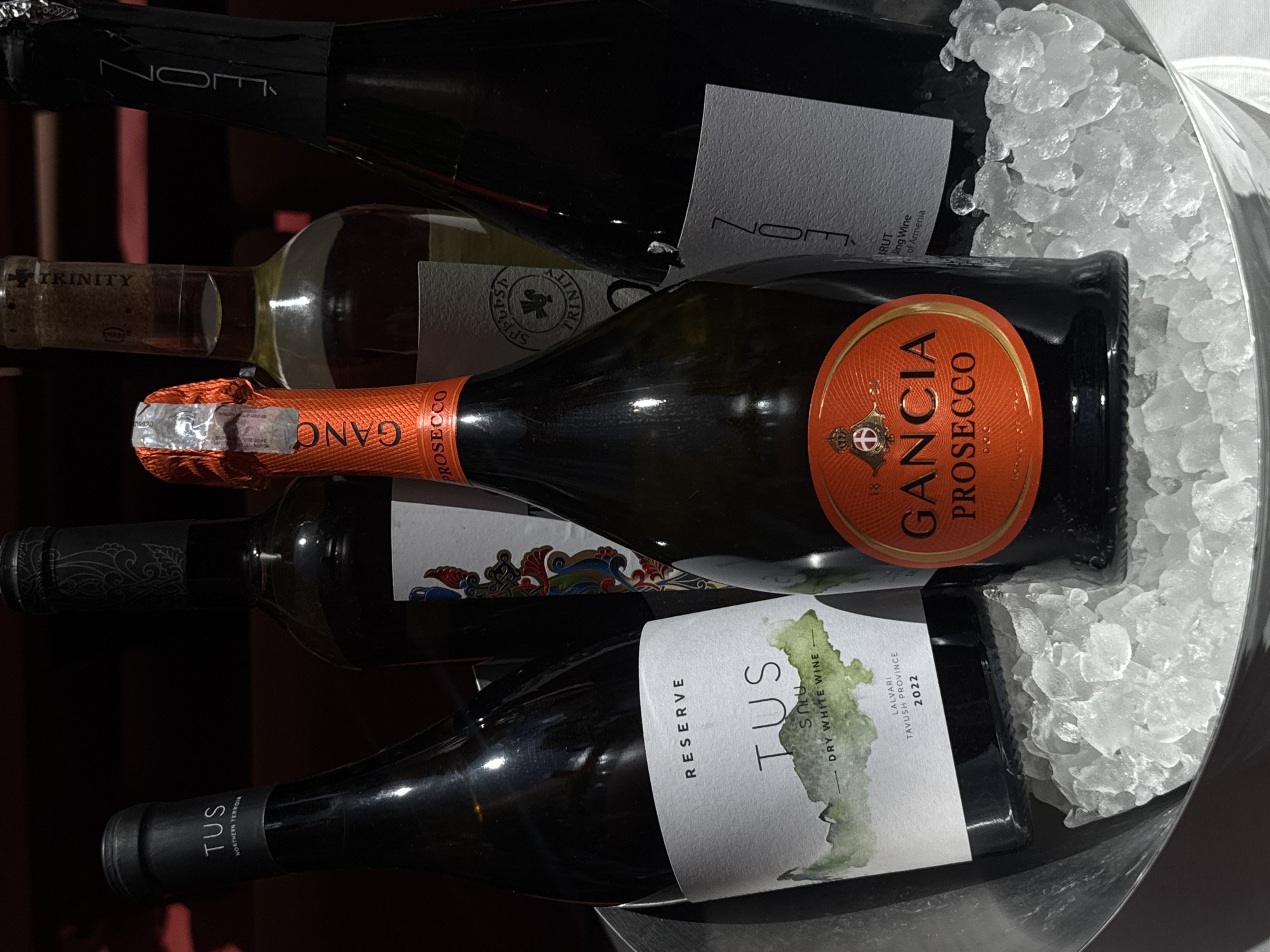 The image shows several bottles of wine in an ice bucket, likely at a restaurant or bar. Visible brands include Gancia Prosecco, TUS Dry White Wine from Lalvari, Tavush Province (2022), and a bottle with "Trinity" visible on the label. Another bottle has the label "NOM". Given the mention of "Wine of Armenia" and the location of Tavush Province, the scene is likely in Yerevan, Armenia. The bottles are surrounded by crushed ice inside a shiny black bucket.