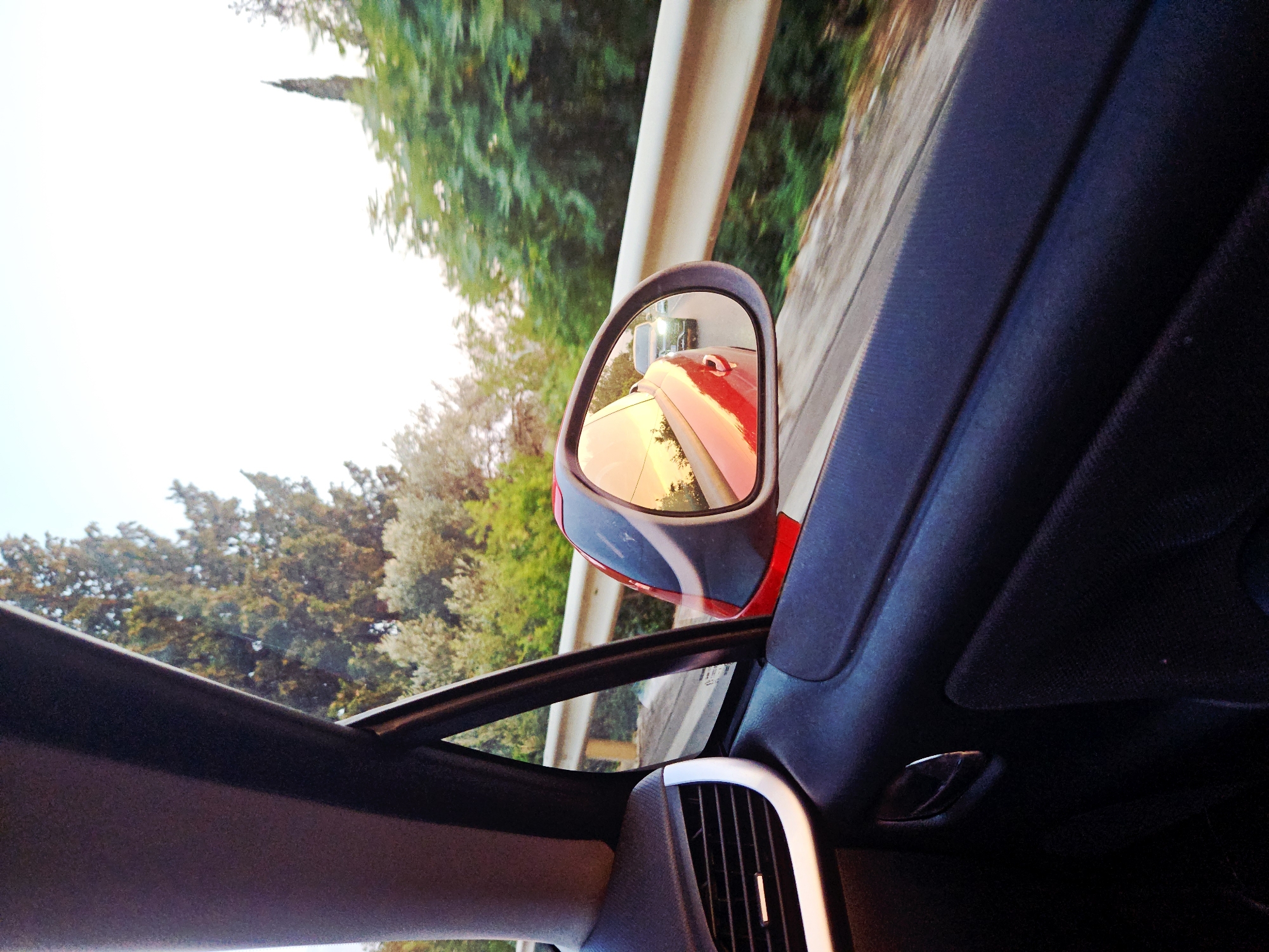 The image is a first-person perspective from inside a car, looking out the side window and into the side mirror. The car is moving along a road, with trees and foliage visible on either side. In the side mirror, another car can be seen following behind. The location is Krstac, Montenegro. The time of day appears to be late afternoon or early evening, as the light is warm and casts reflections on the car's surface.