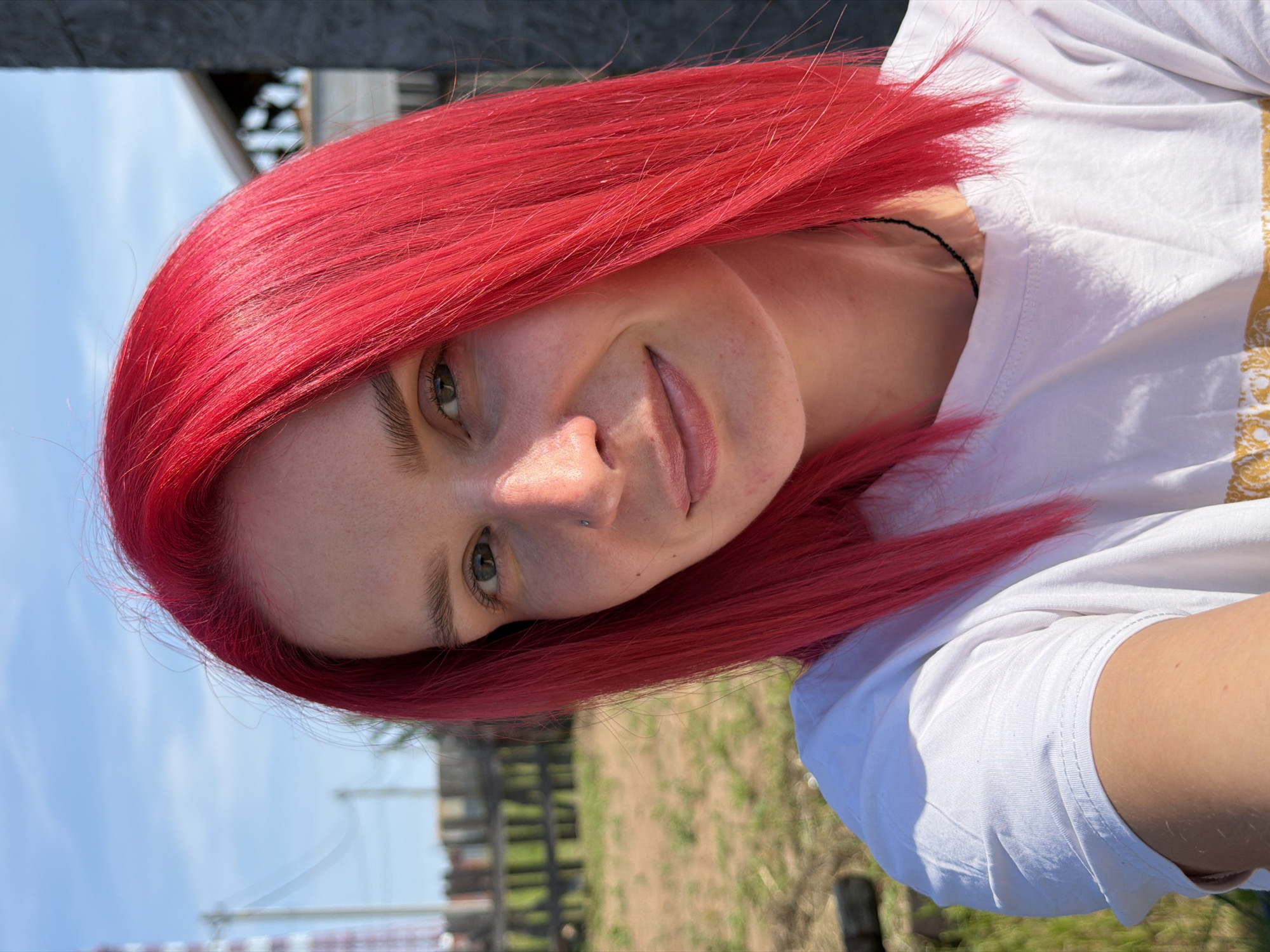 A bright, eye-level, medium shot shows a young woman with vibrant red hair, facing forward and smiling slightly. She is wearing a white t-shirt and a thin black necklace. Her right side is illuminated by sunlight, creating a visible shadow on the left side of her face. A small nose ring is visible on her left nostril. In the background, a blurred outdoor scene includes a wooden fence, dry grass, and a tall, industrial-looking structure with a white top. The sky above is a clear blue with a few wispy clouds. The overall lighting suggests it is a sunny day. The presence of the industrial structure and the style of housing visible in the background suggest a location in a more rural or industrial area of a city, consistent with some parts of Chaikovsky, Russia.