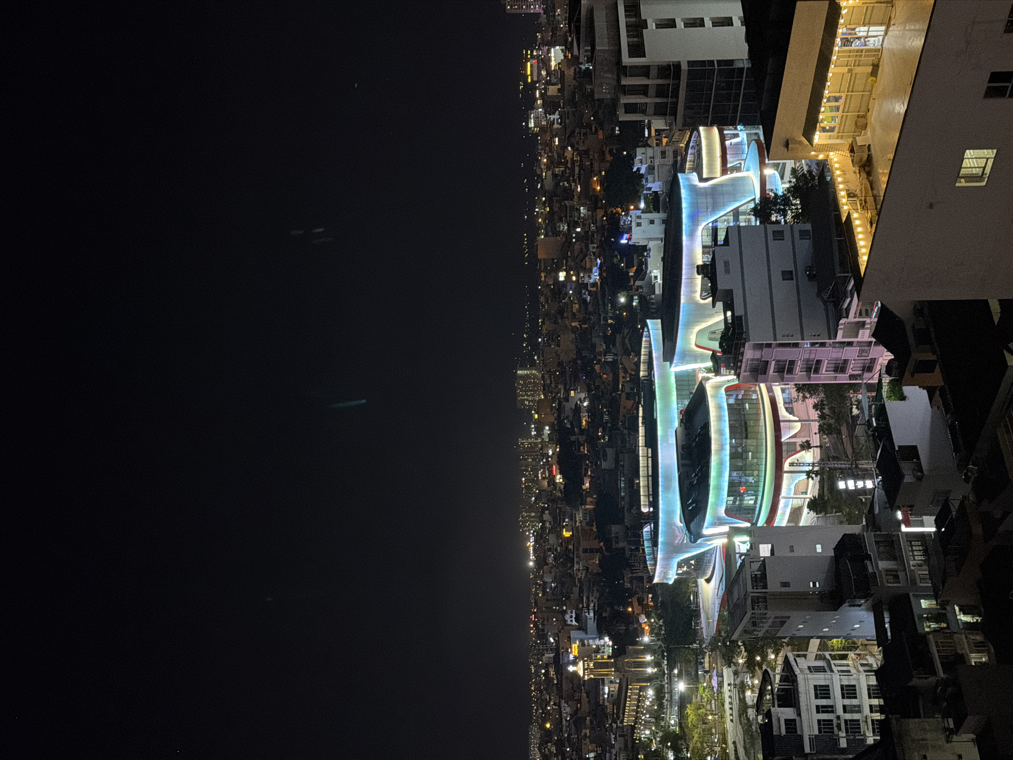 An elevated, night-time view captures a cityscape in Nha Trang Ward, Vietnam, dominated by modern architecture and illuminated by artificial lights. In the foreground, the top of a building with a rooftop patio adorned with string lights is visible. Further into the scene, a strikingly contemporary building with a curved, multi-colored illuminated facade stands out. To the left of this structure, a white building with multiple stories and a distinctive stepped design is present. In the mid-ground, a large, illuminated building with a circular design and a ramp-like structure is prominent. Behind these central elements, a dense urban landscape unfolds, characterized by numerous buildings of varying heights and styles, all lit up against the dark sky. A church with illuminated spires and a prominent cross is situated to the far left of the mid-ground. The distant horizon is dotted with the lights of more buildings, indicating the vastness of the city. The sky is dark and clear, with faint, streak-like anomalies, possibly lens flares or atmospheric effects, visible in the upper portion of the image. No individuals are discernible in the scene.