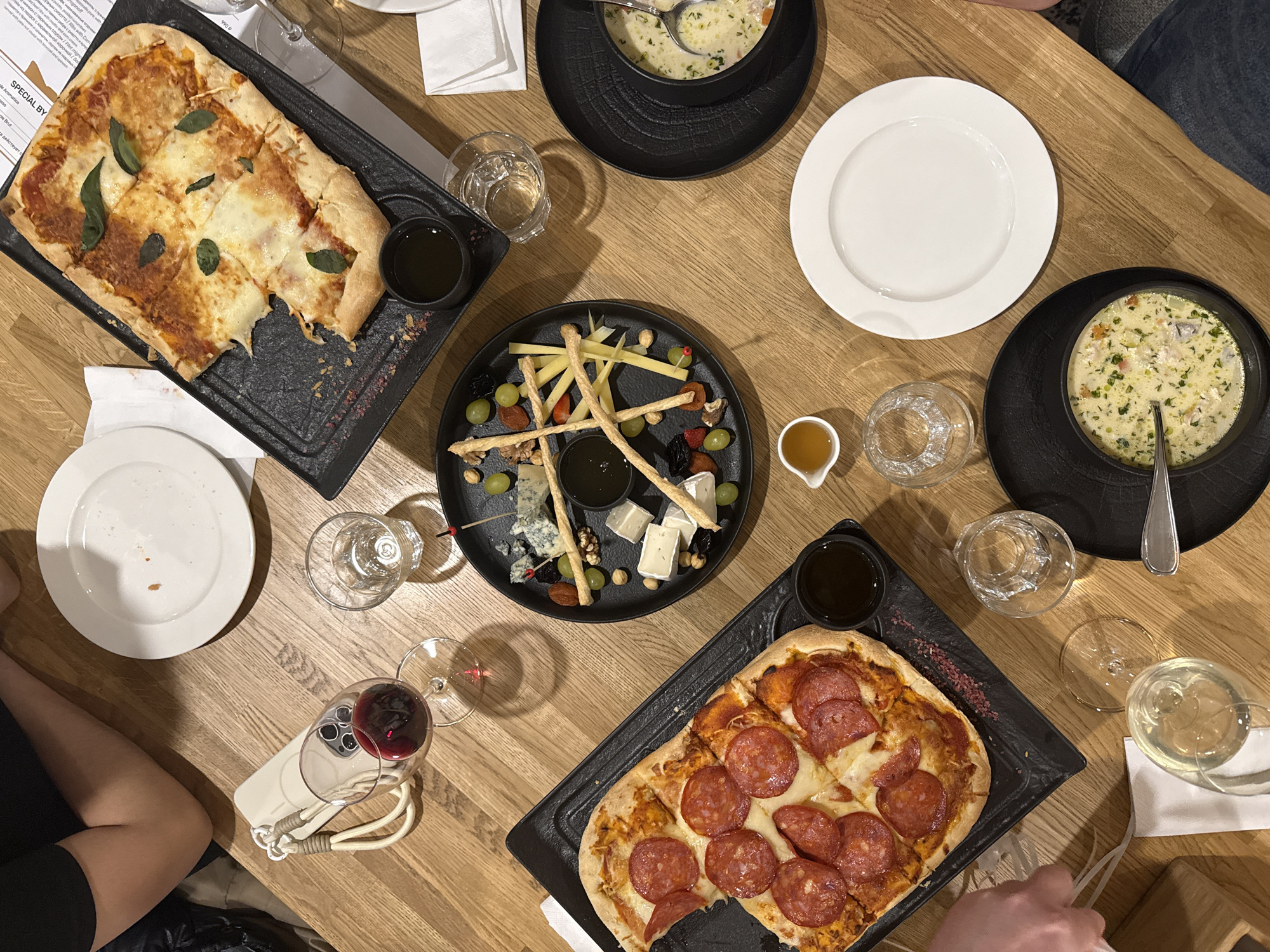 The image depicts a tabletop setting with a variety of dishes. There are two pizzas, one with pepperoni and one with cheese and basil, served on black rectangular platters. A cheese plate with various cheeses, grapes, nuts, and dried fruits is also present, along with two bowls of soup served in black bowls, each with a spoon. Several drinking glasses, some filled with clear liquids, and one filled with red wine, are scattered around the table. White napkins are positioned near some of the glasses. A white cell phone with a wine glass resting on it is on the lower left side of the table. The tabletop is made of light-colored wood. It is not possible to determine the location in Moscow from the given image. There is interaction of people at the table, with body parts such as an arm and a hand visible.