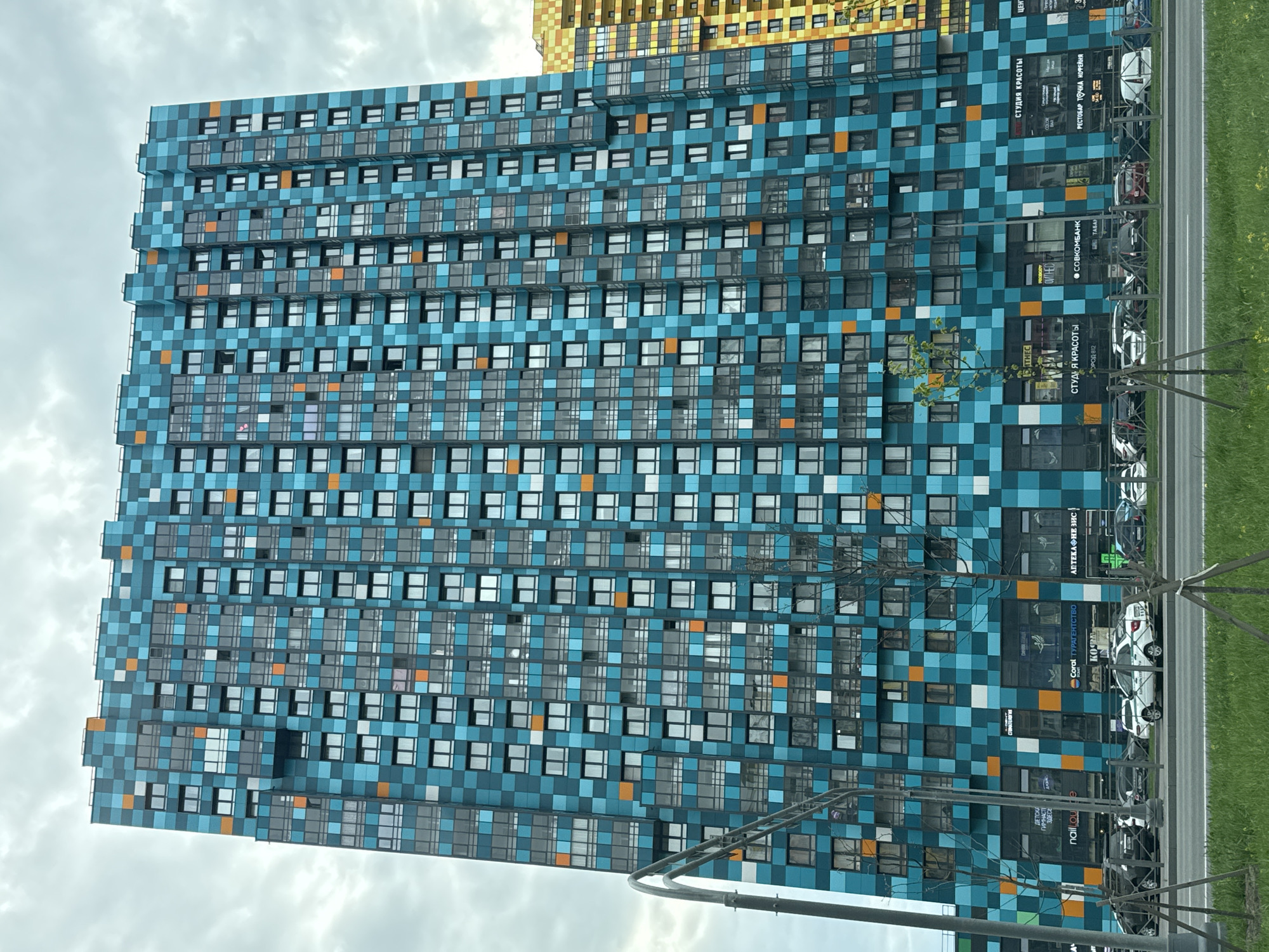The image shows a tall, modern apartment building in Saint Petersburg, Russia. The building has a distinctive facade decorated with a mosaic of blue, teal, and white tiles, with occasional orange squares. Rows of windows are visible across the building's surface. The lower part of the building has retail spaces with storefront signs written in Russian. Cars are parked along the road in front of the building. A sparse tree is in the foreground, with thin branches and small leaves. The sky is overcast and cloudy. A neighboring building with a gold façade can be seen in the background, near the top of the image. A gray metal structure is also visible, partly obstructing the building. The image is taken from a low angle, looking upwards at the building.